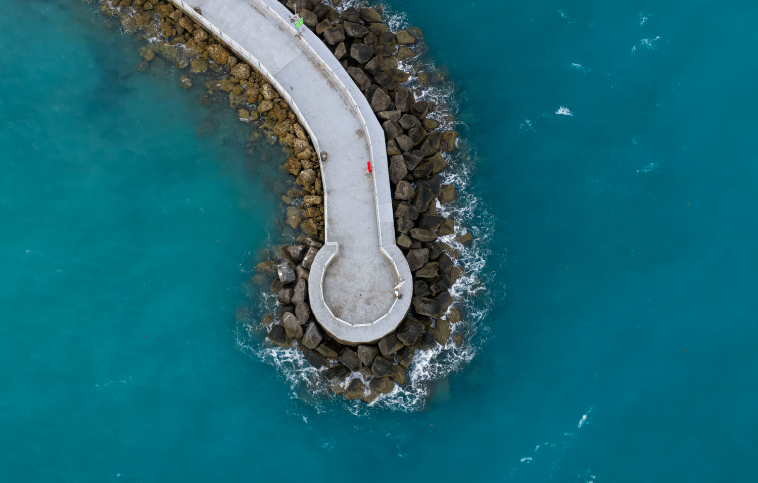 Aerial view of a curved concrete pier surrounded by turquoise ocean waves with a lone person in red walking along it.