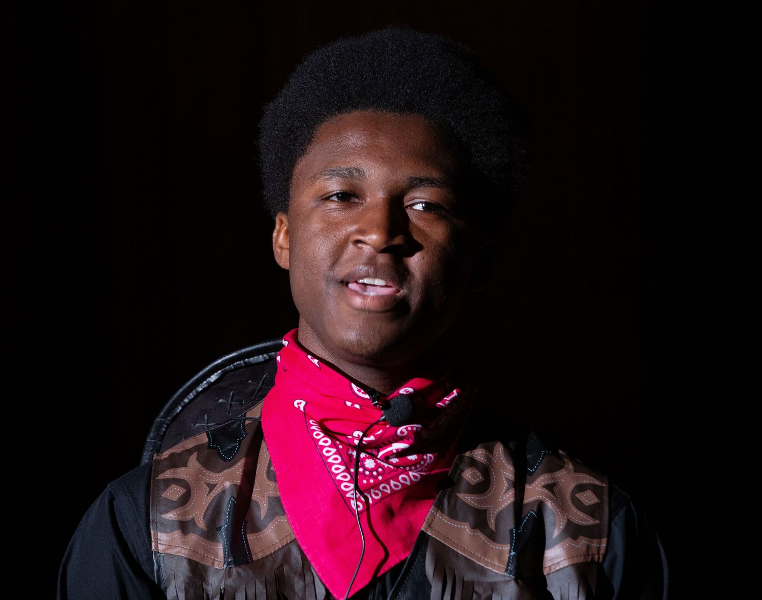 Person of color with pink bandana poses against black background in dramatic theater lighting.