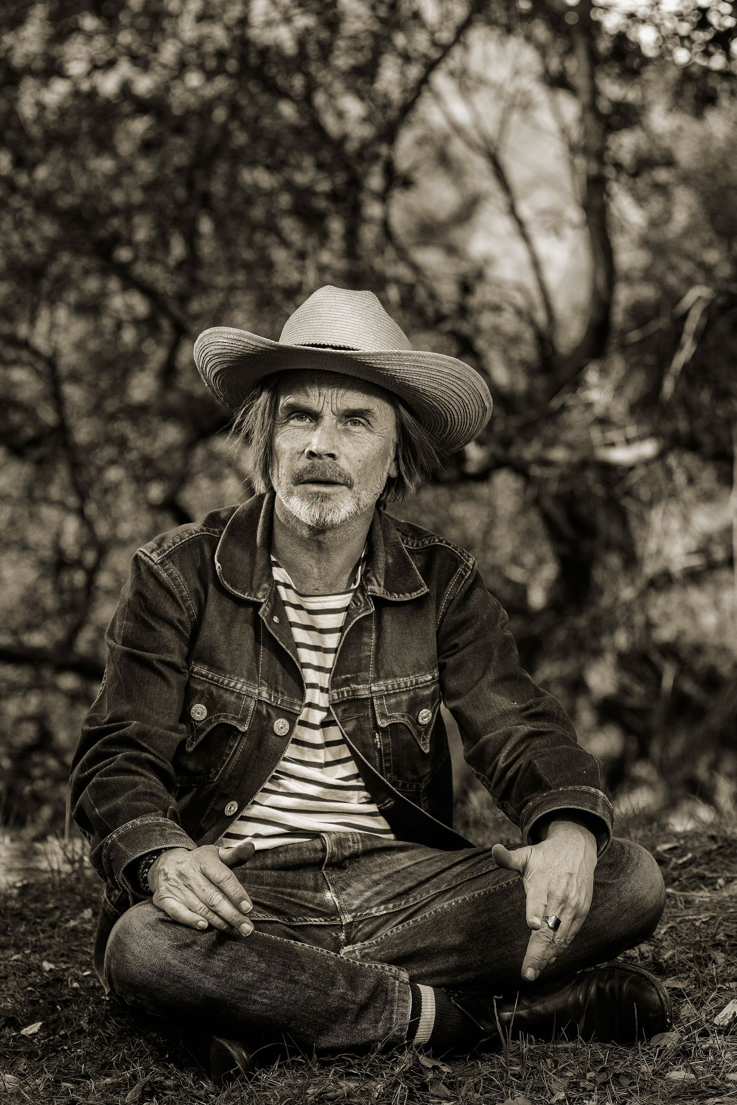 Black and white portrait of a rugged man in cowboy hat and denim jacket sitting cross-legged outdoors.