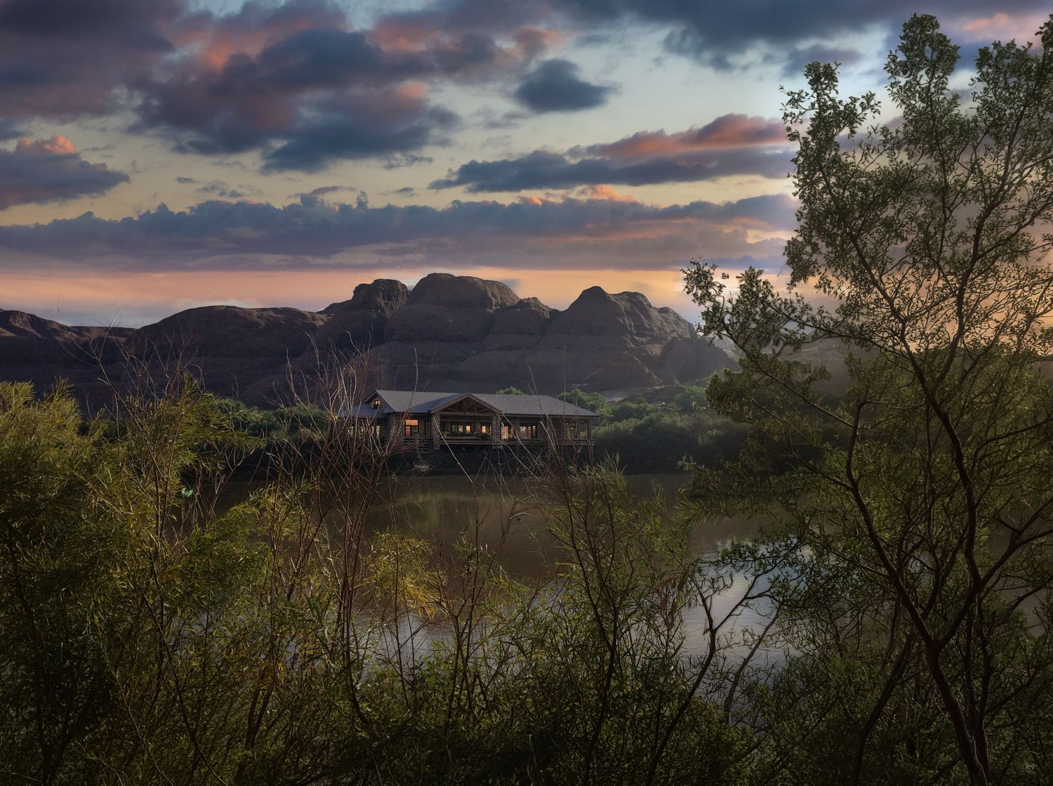 Scenic desert landscape at sunset with a pavilion reflecting on a calm river, surrounded by lush trees and rocky mountains.