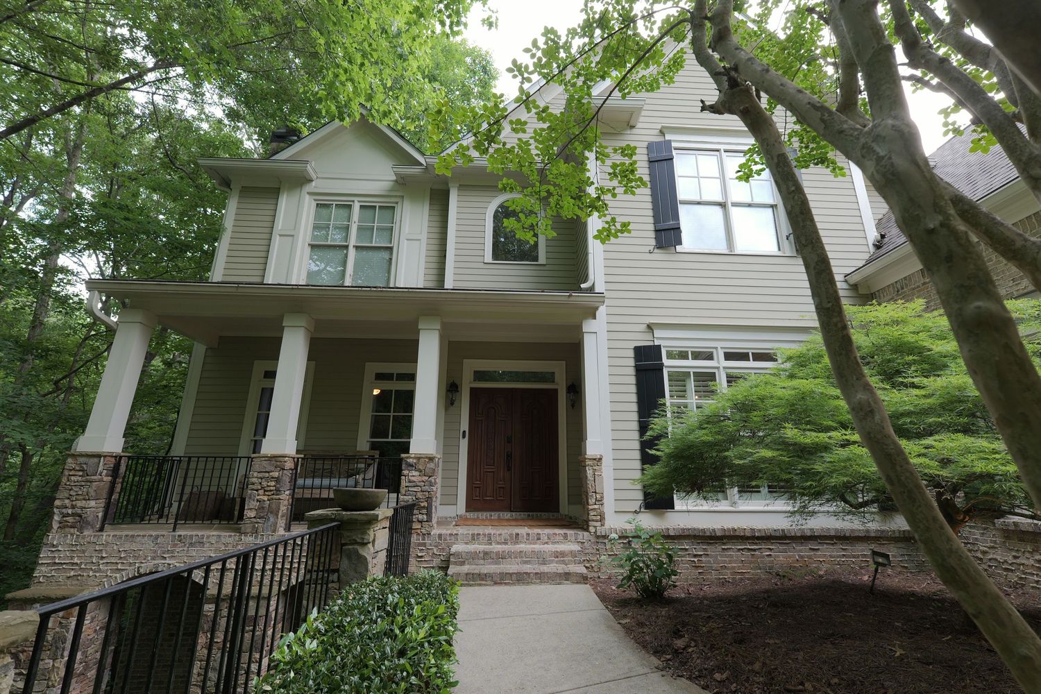 Two-story craftsman home with stone accents, white columns, dark shutters, and lush surrounding trees.