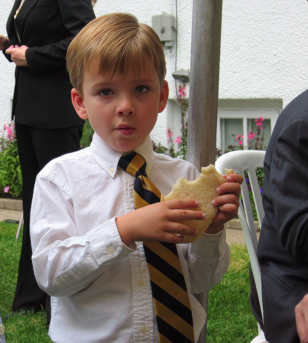 A child in a white shirt and striped tie takes a bite of food while standing outdoors.