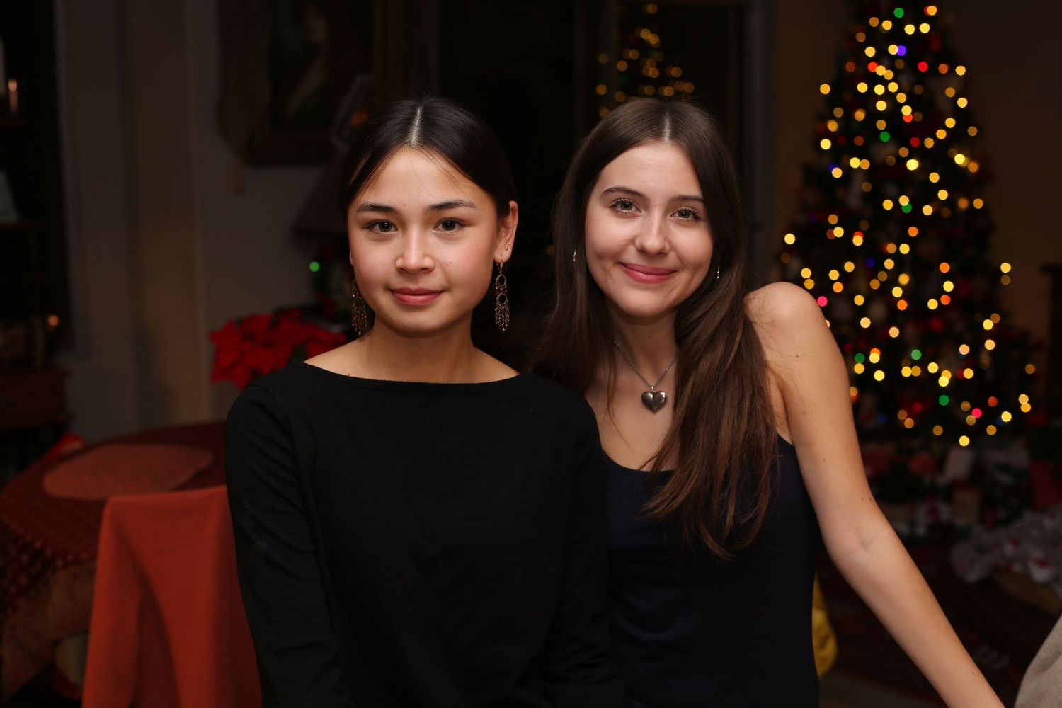 Two young women in black outfits smiling together near a decorated Christmas tree at a holiday gathering.