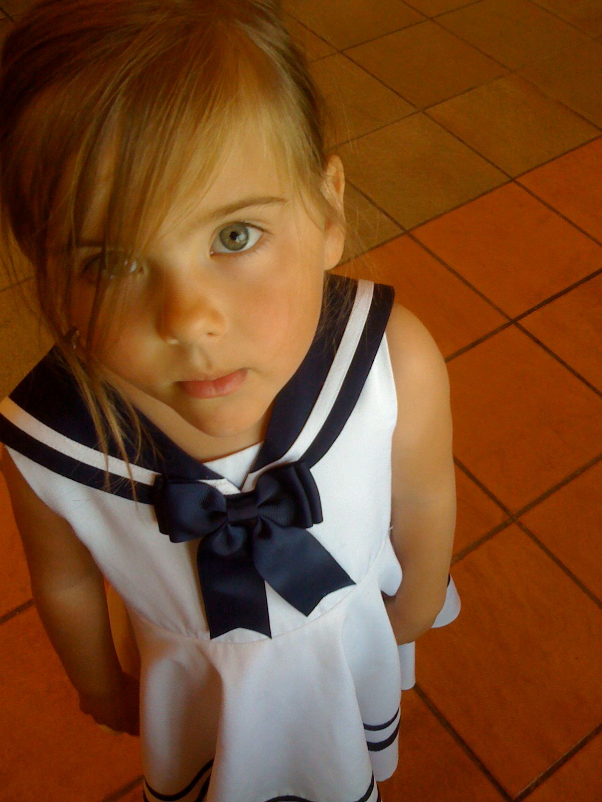 Young person in white sailor-style dress with navy blue bow against terracotta tile floor.