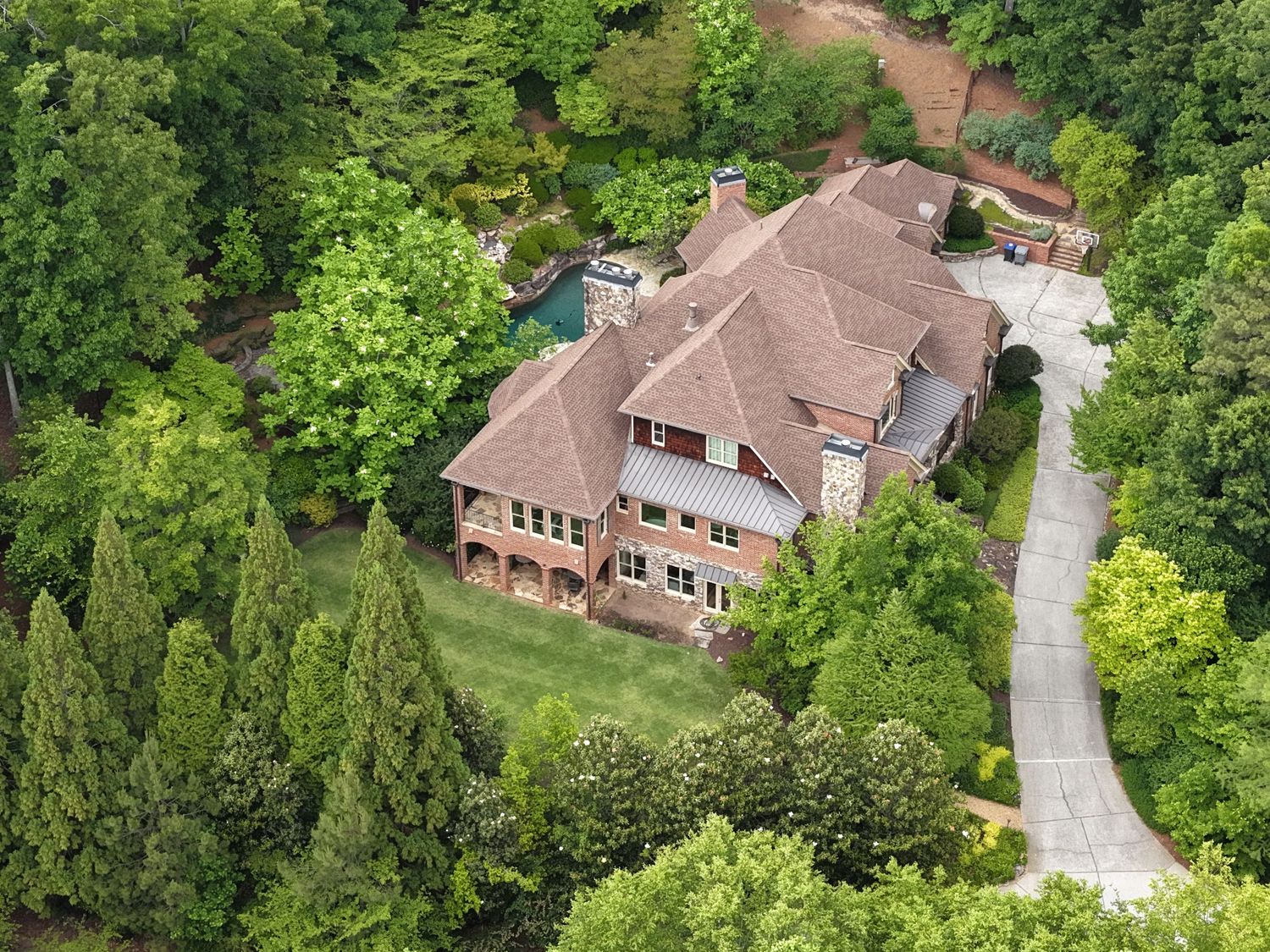 Aerial view of a large brick estate home surrounded by lush green trees with a curved driveway and pool.