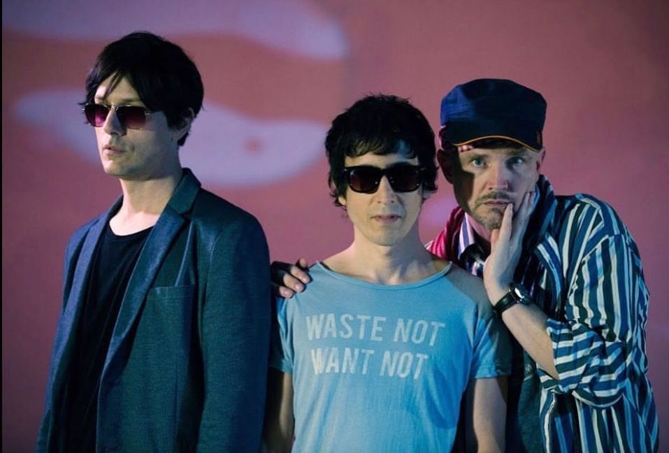 Three musicians in sunglasses posing against pink backdrop in retro indie band photo.