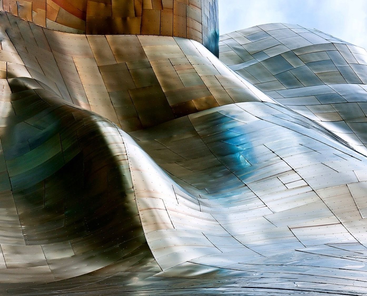 Abstract curved metallic panels of a modern building reflect blue and gold tones under a cloudy sky.