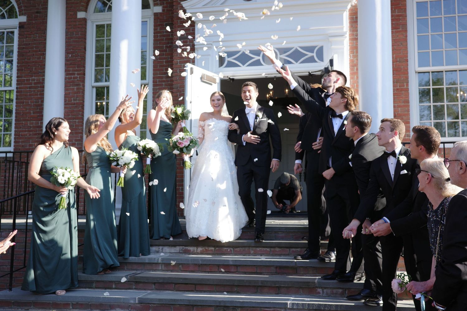 Wedding party stands on church steps as flower petals are tossed during a joyous celebration.