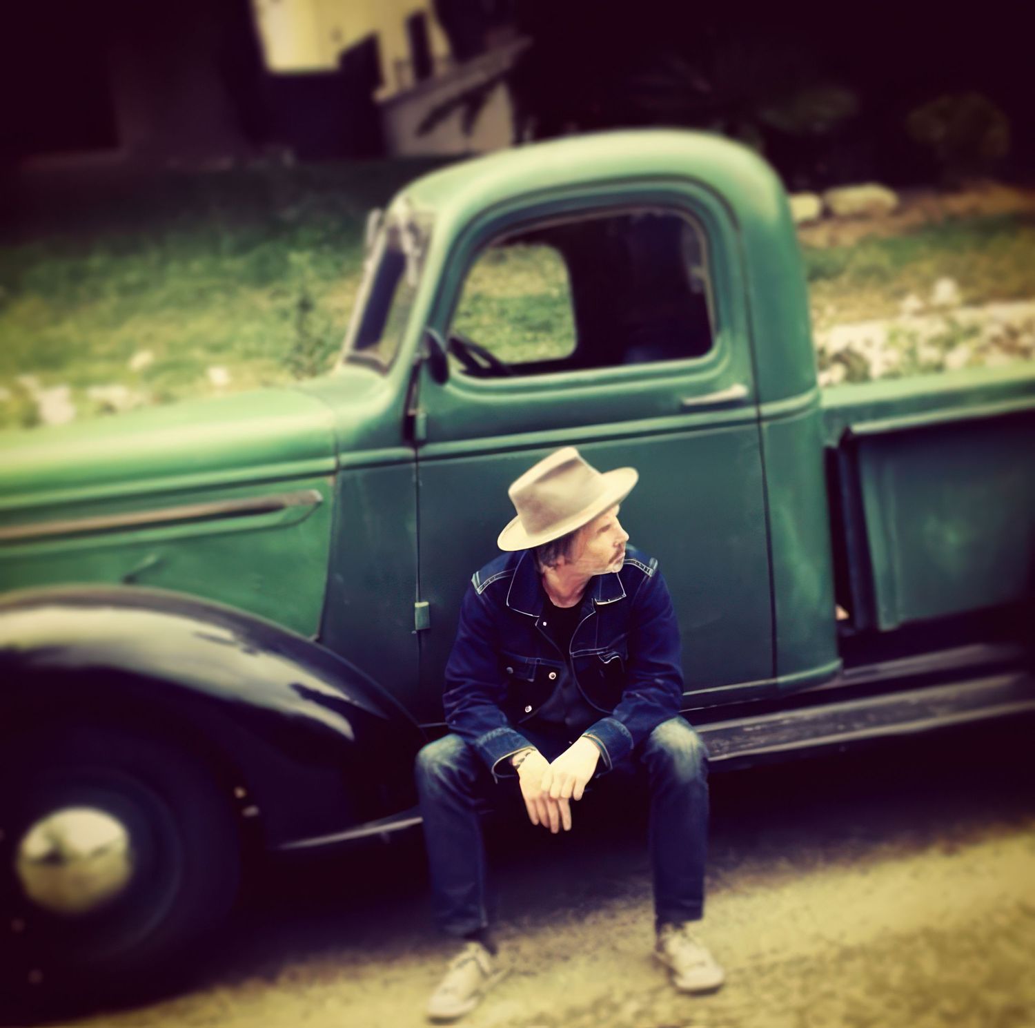Vintage green pickup truck parked on a dirt road with someone sitting casually beside it.