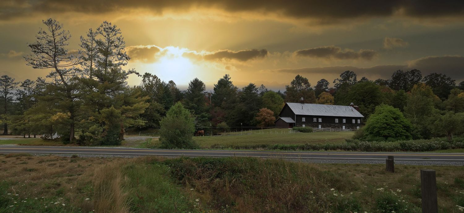 Rural farmhouse at sunset with dramatic golden sky, lush green trees, and a quiet country road in a scenic landscape.