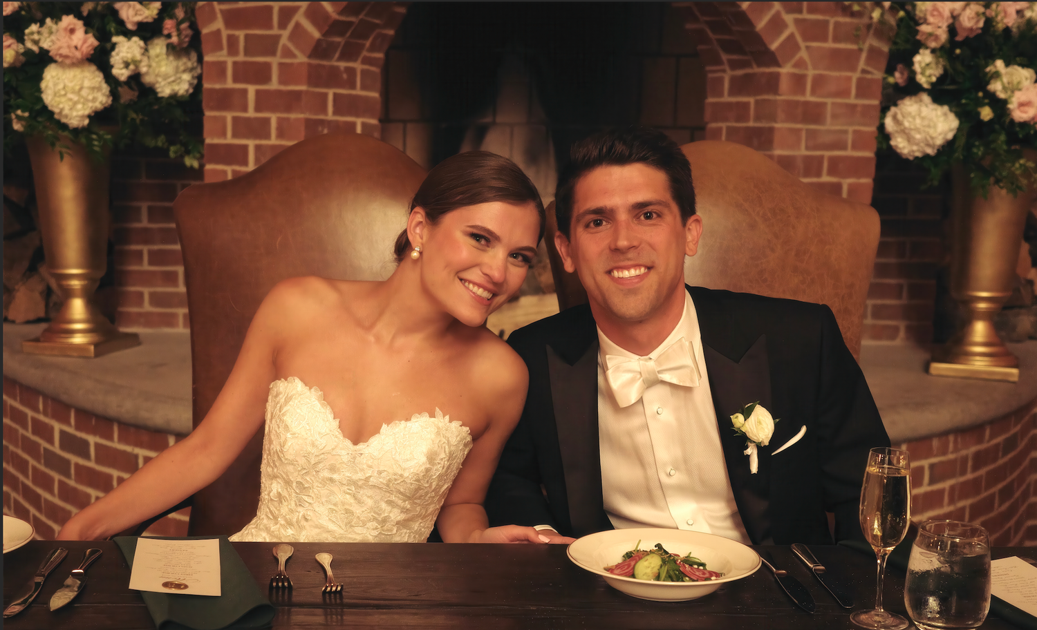 A couple sits together at their wedding reception table with brick archway and floral decorations in the background.