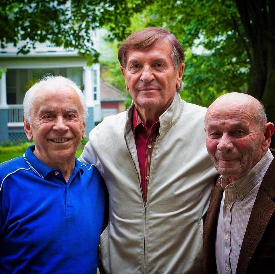 Three senior men smiling together outdoors in a garden setting.