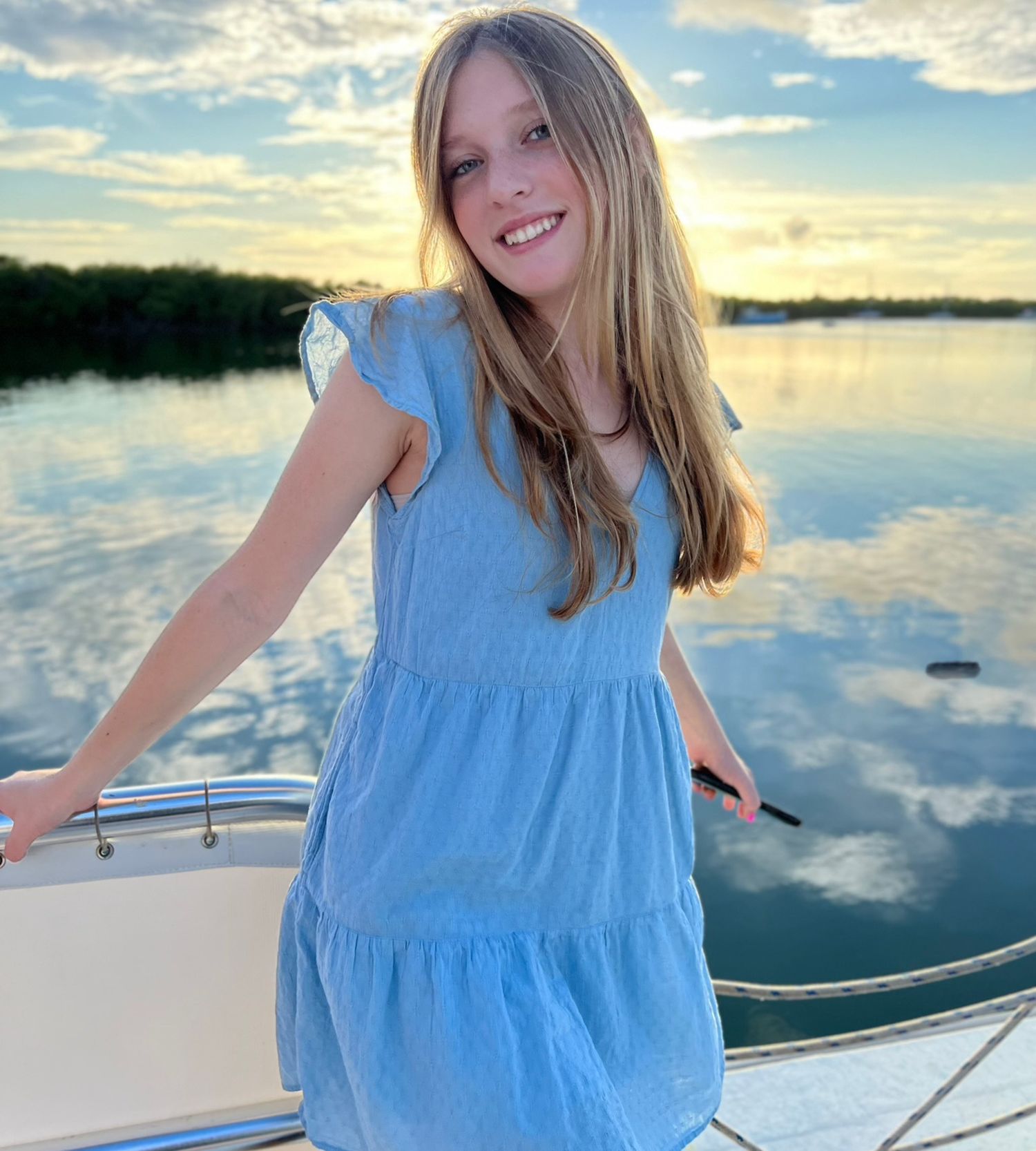 A person in a blue dress poses on a boat during sunset with reflective water in the background.