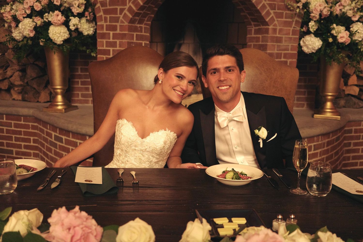 Wedding reception dinner setup with floral centerpieces and brick wall backdrop.