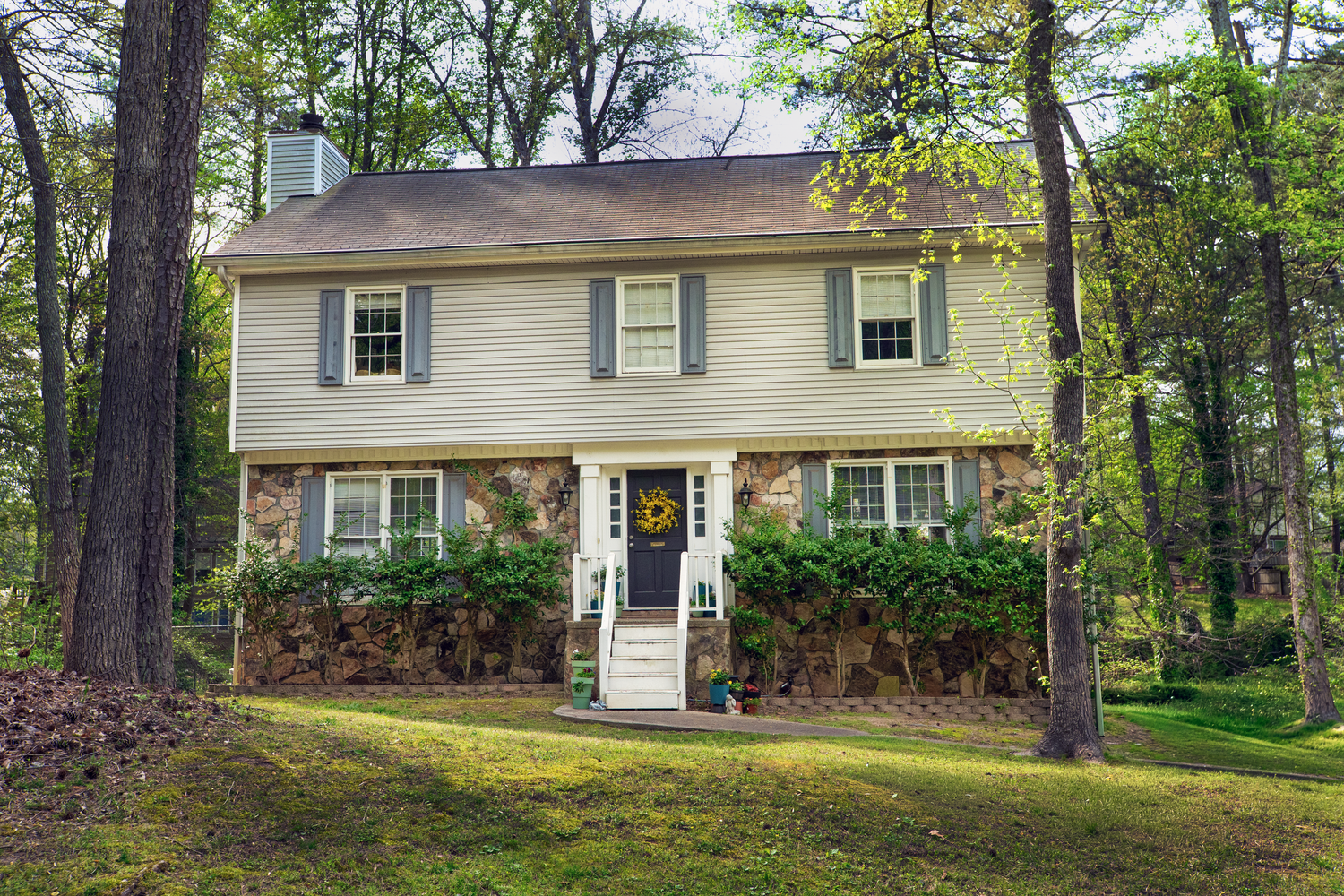 Two-story colonial home with stone facade, gray shutters, and lush green trees surrounding the property.