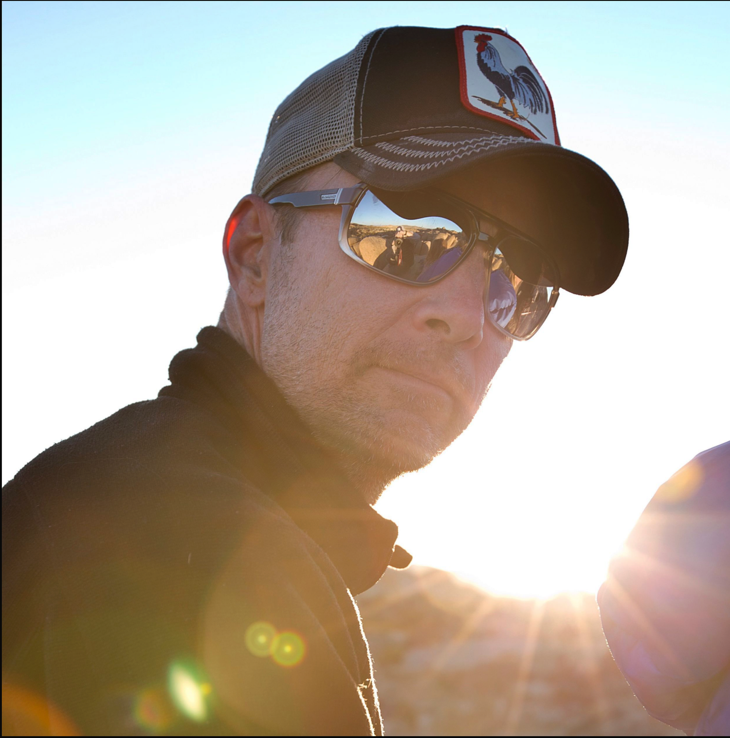 Silhouetted person wearing sunglasses and baseball cap in golden sunlight with lens flare effects.
