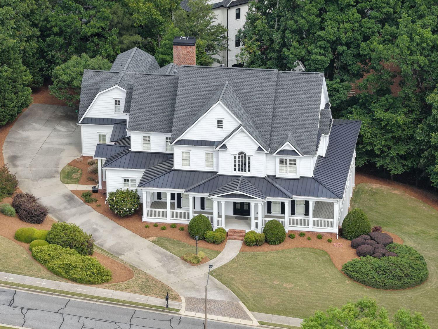Aerial view of a large white two-story home with gray roof, wraparound porch, and circular driveway surrounded by lush trees.