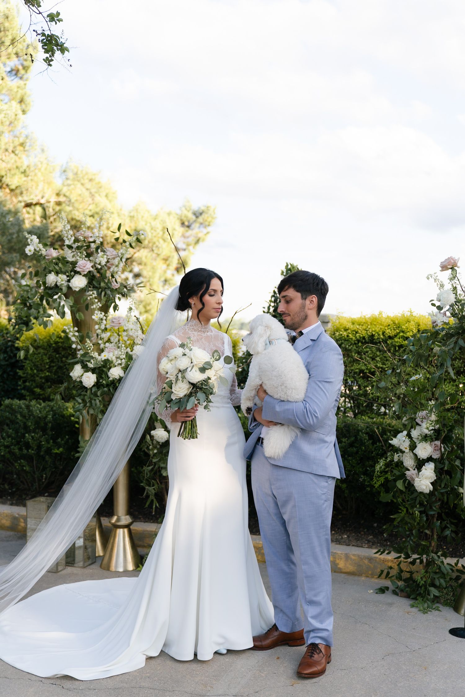 Newlyweds share romantic moments in garden setting with white floral arrangements and flowing bridal veil.