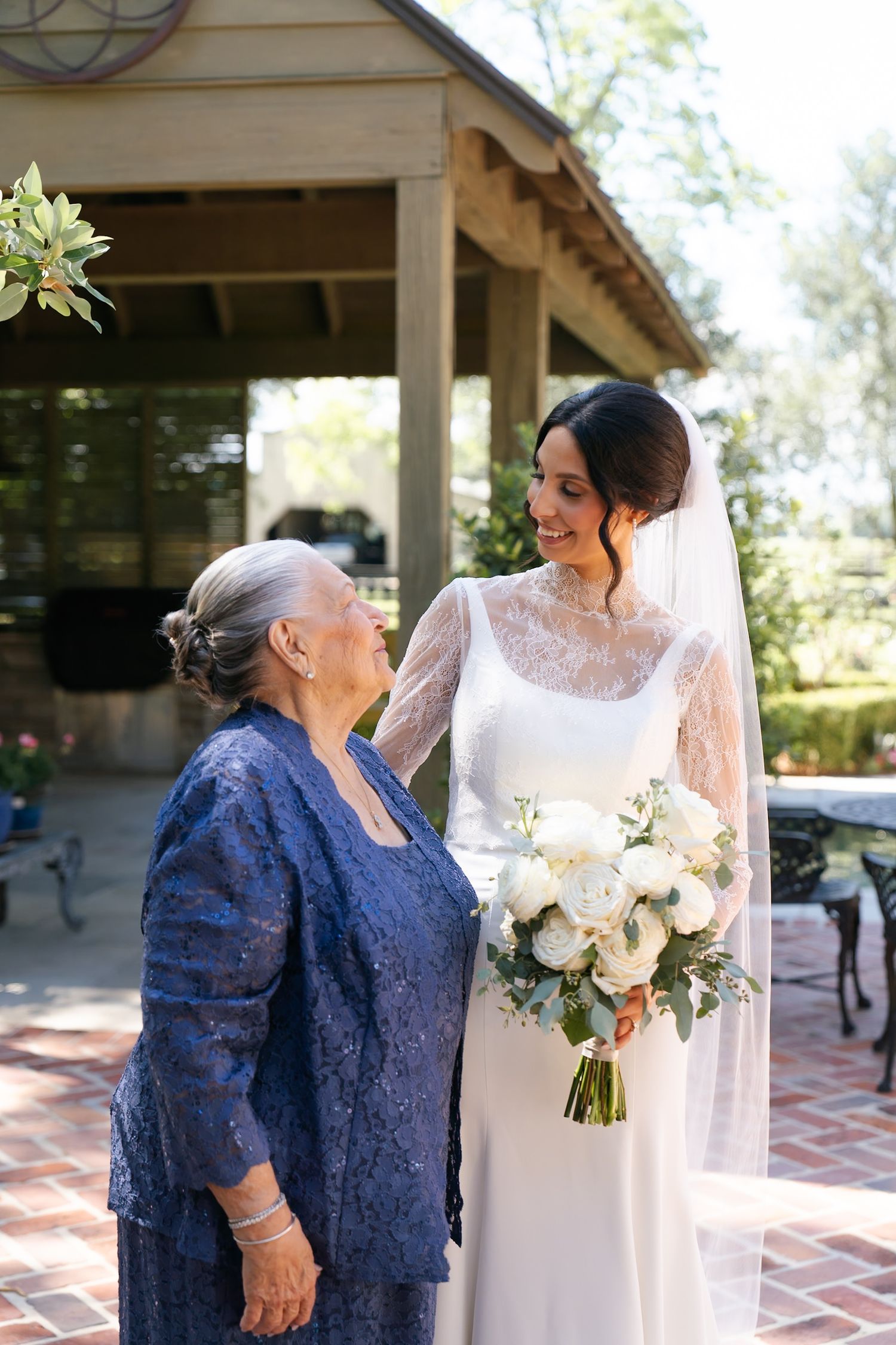 Bride in white lace dress shares emotional moment with family member in navy blue at outdoor venue.