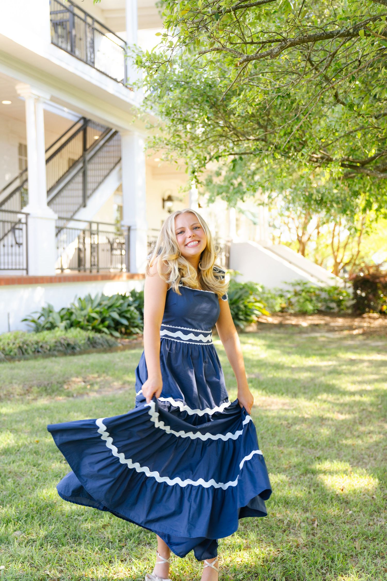 A person in a navy blue dress with white trim twirls in a grassy yard near a white house.