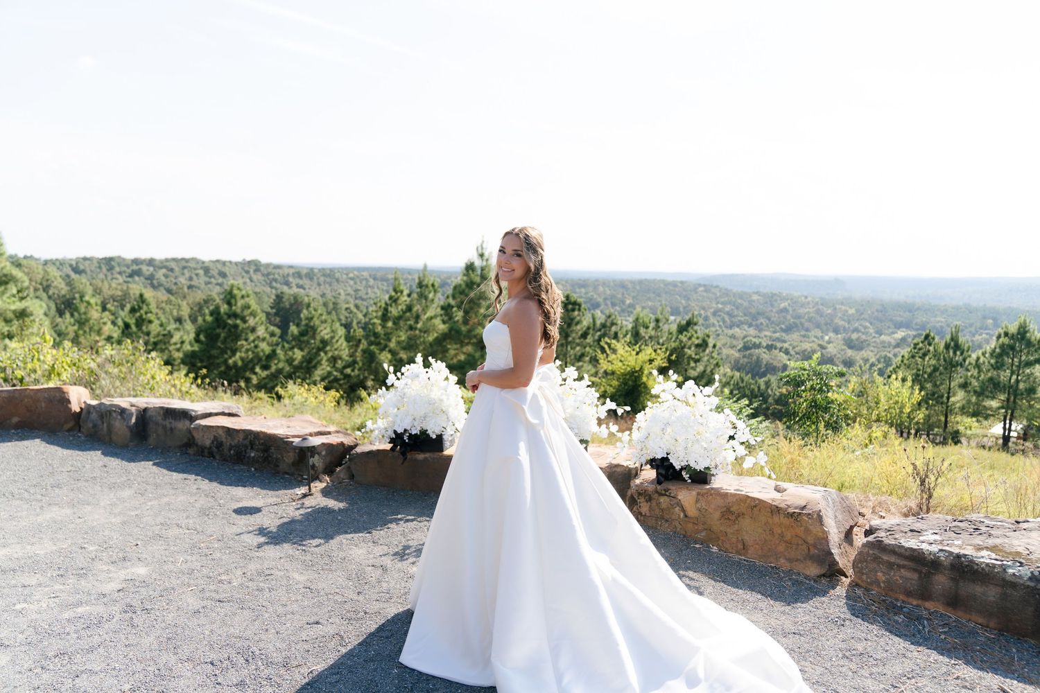 A bride in a white wedding dress stands at a scenic mountain overlook on a sunny day.