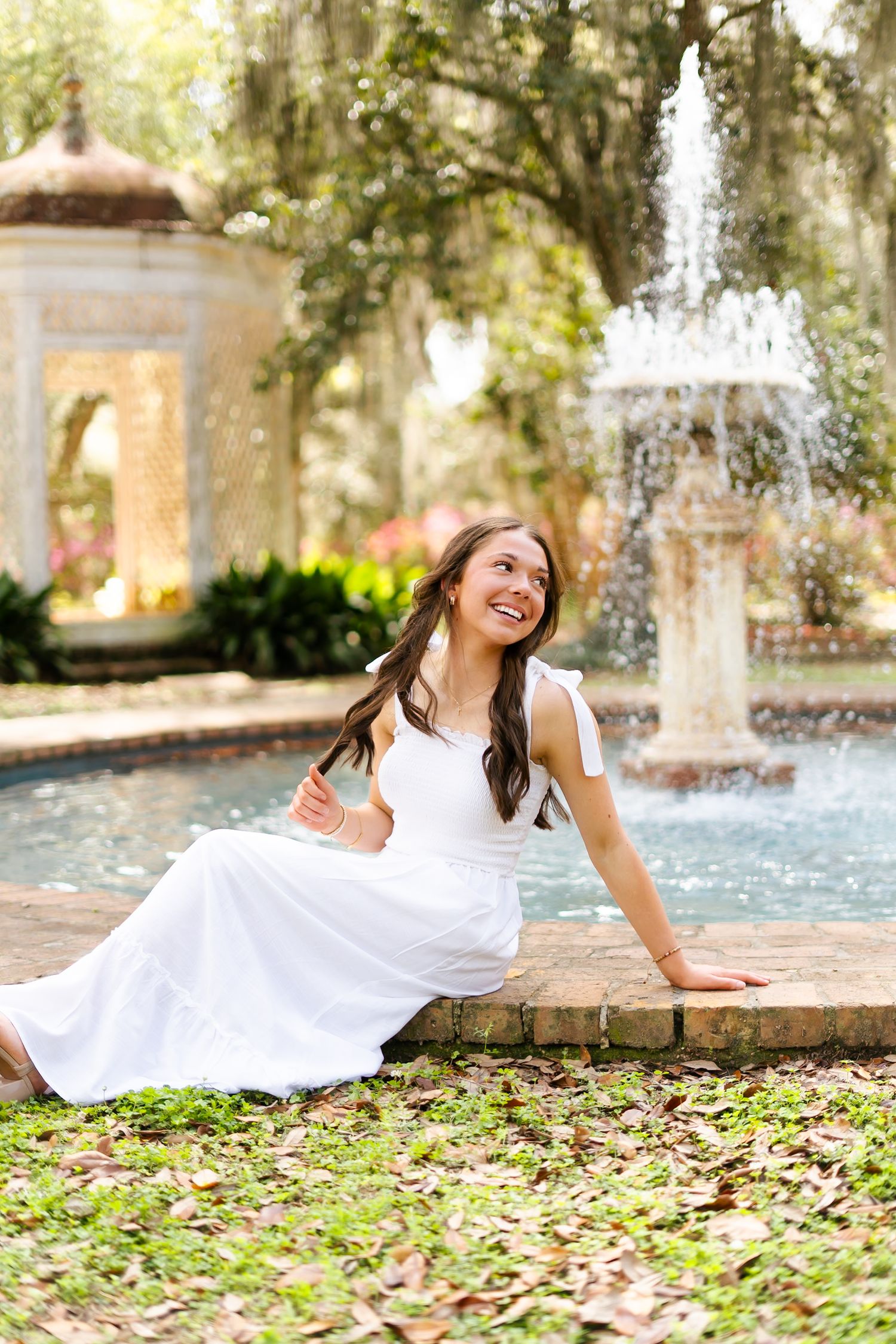 Person in white dress lying gracefully on grass with autumn leaves and fountain in background at garden setting.