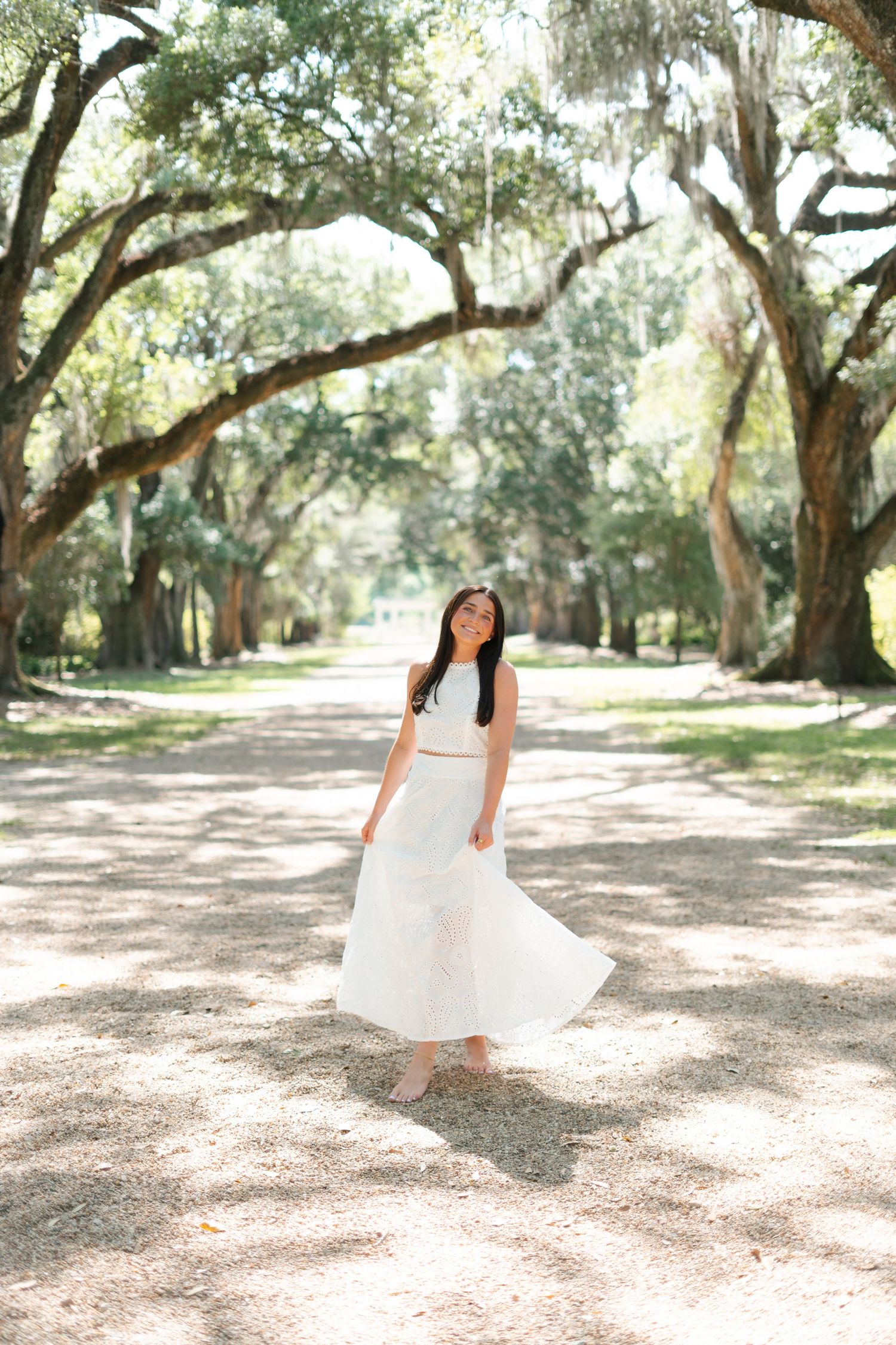 Person in white dress twirls under moss-covered oak trees in dreamy outdoor setting.