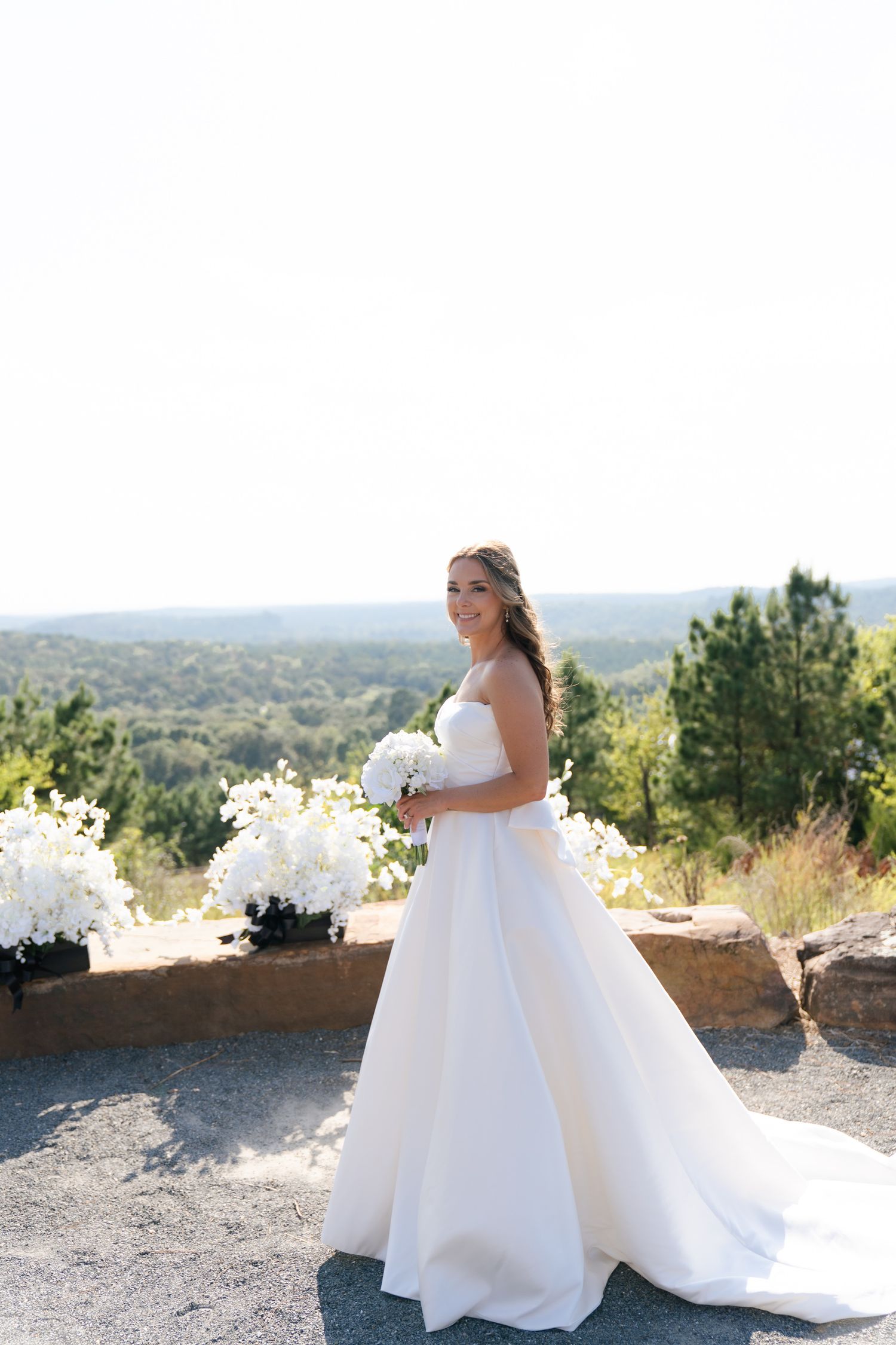 A bride in a flowing white wedding dress poses against a scenic mountain overlook on her special day.