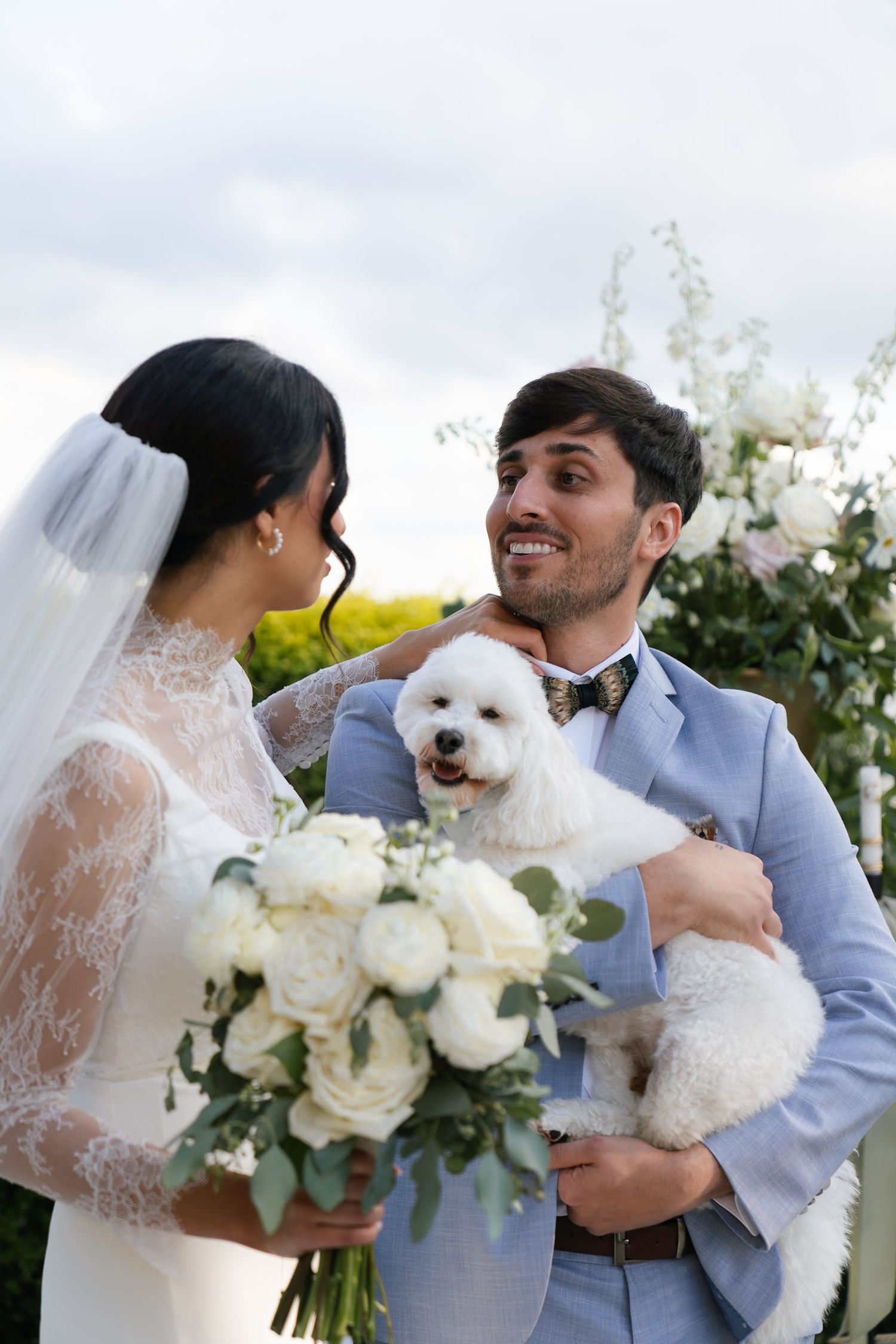 Newlyweds share joyful moment with small white dog while holding white rose bouquet.