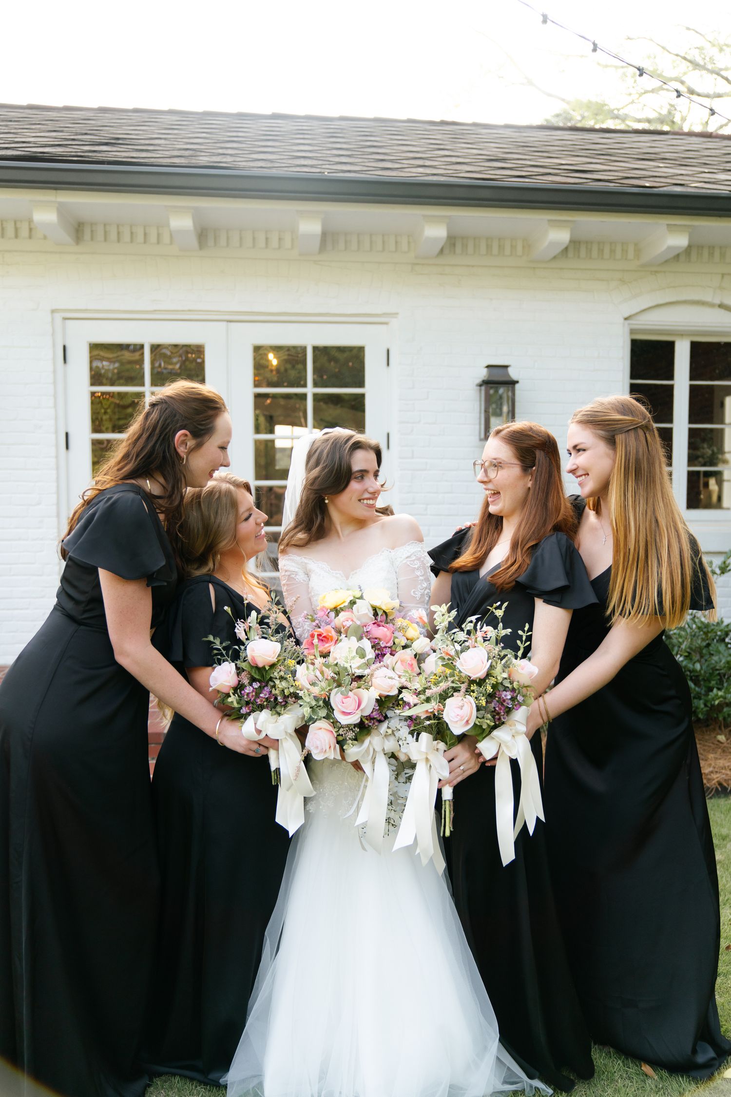 Bridesmaids in black dresses share joyful moment with bride in white gown outside.