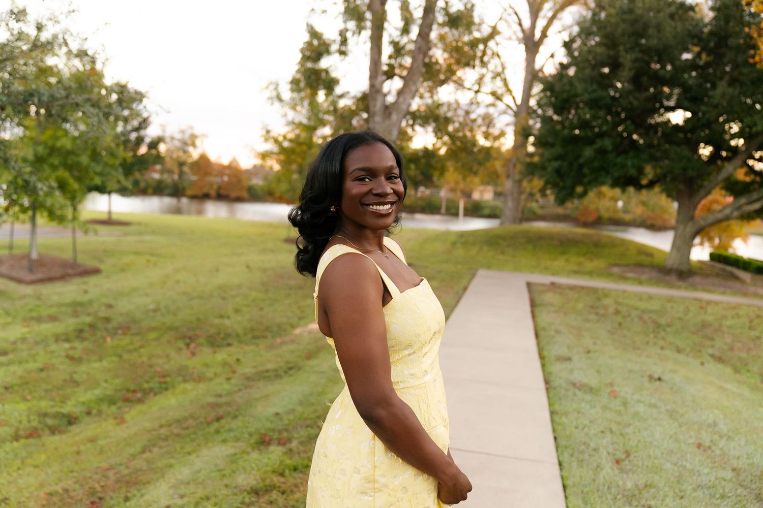 A woman in a yellow dress poses outdoors during golden hour on a path surrounded by green grass and trees.