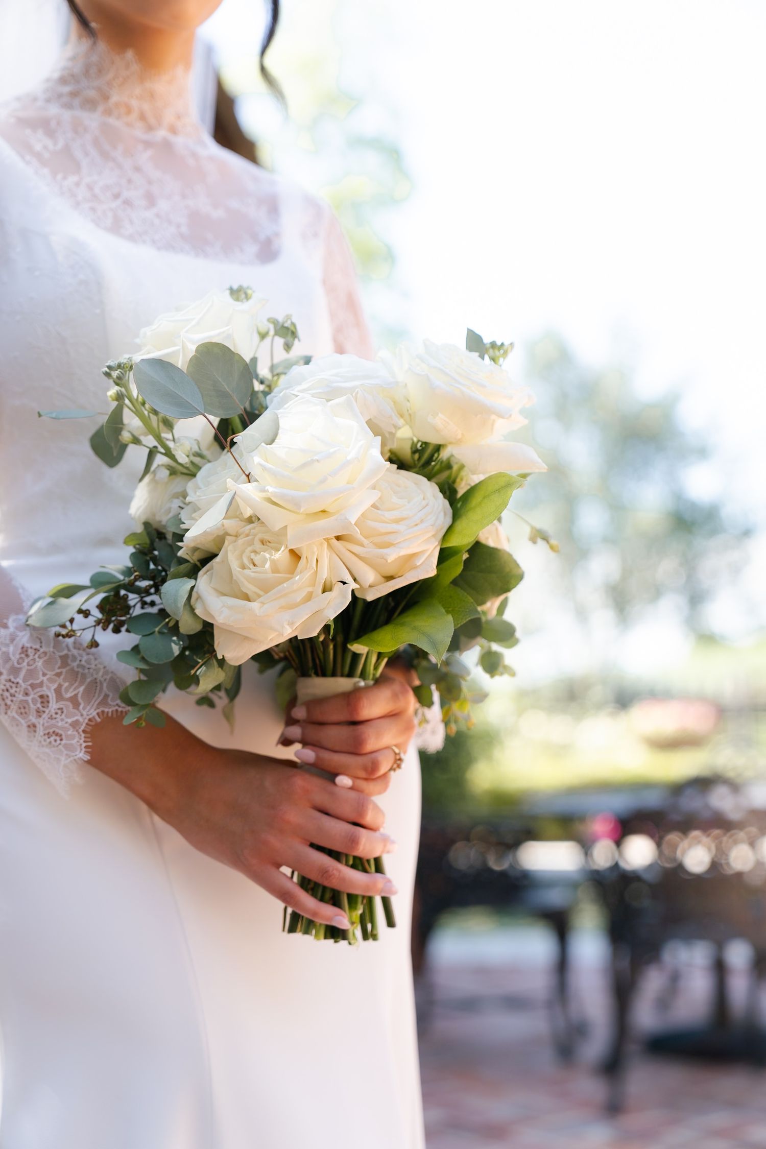 Close-up shots of white rose wedding bouquet with eucalyptus held by bride in lace sleeves.