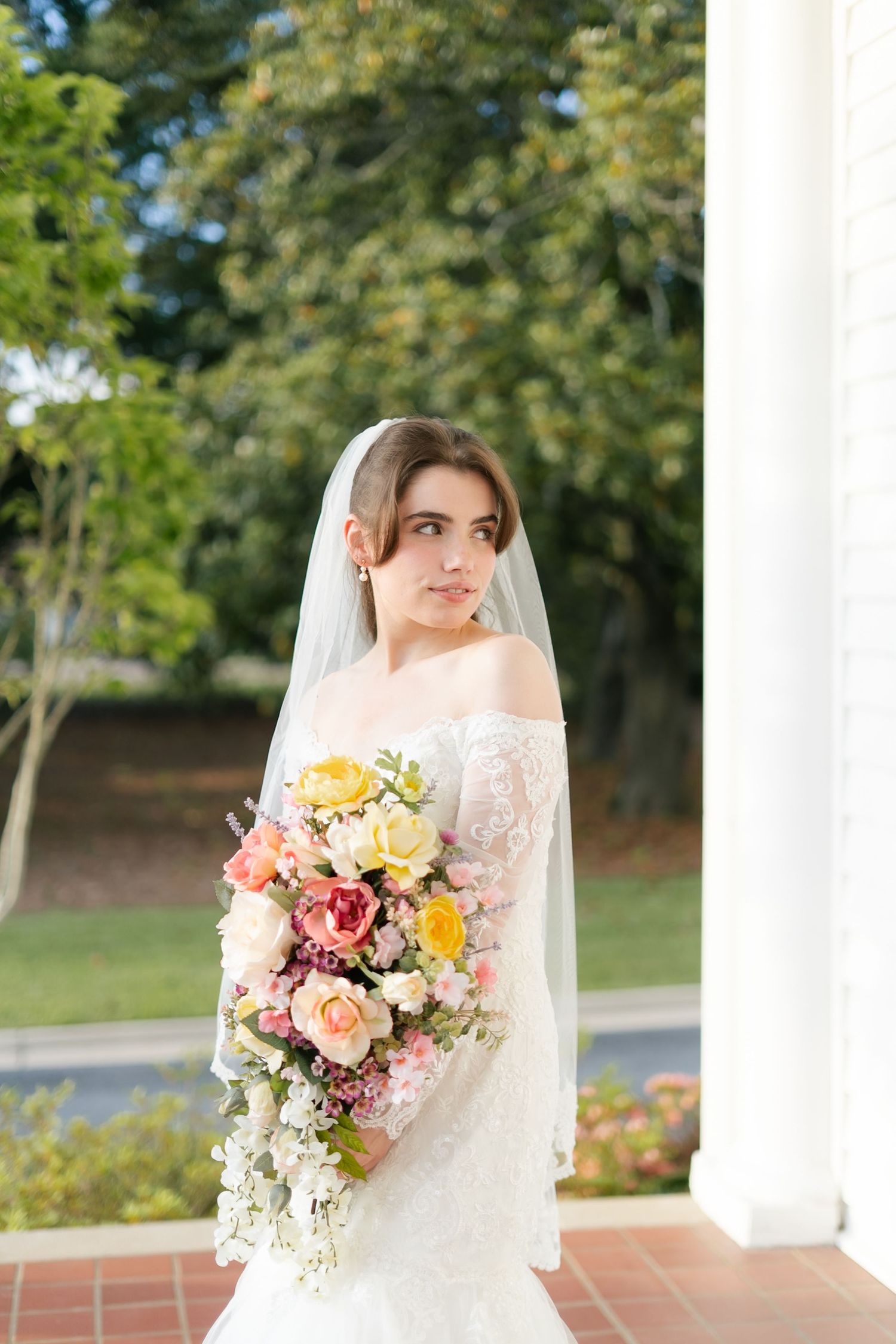 Bride with veil holds colorful bouquet of pink and yellow flowers against white columned backdrop.