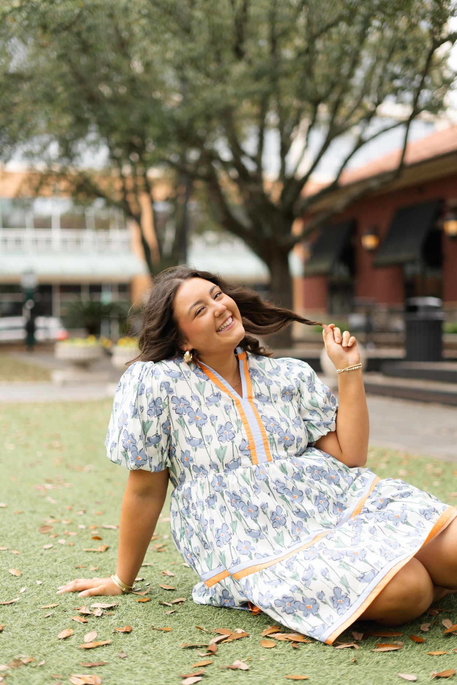 A joyful person in a blue and white floral dress sits on grass near buildings.