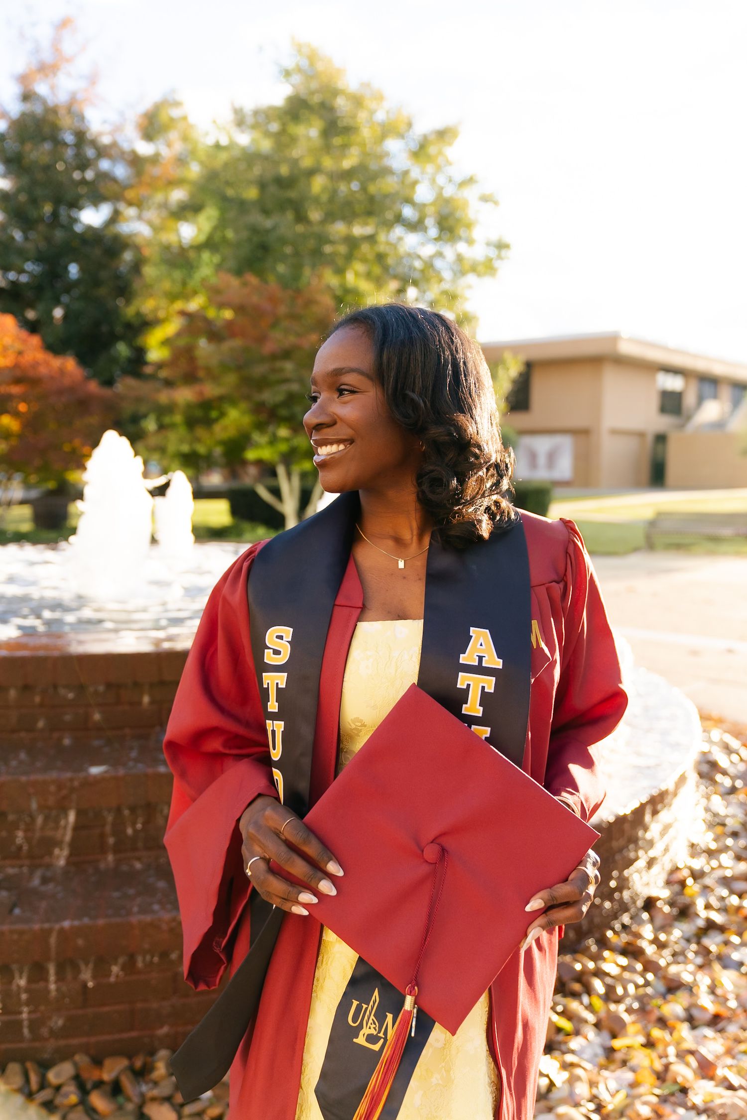 Graduate in red and black academic regalia smiles while holding diploma during autumn graduation photoshoot outdoors.