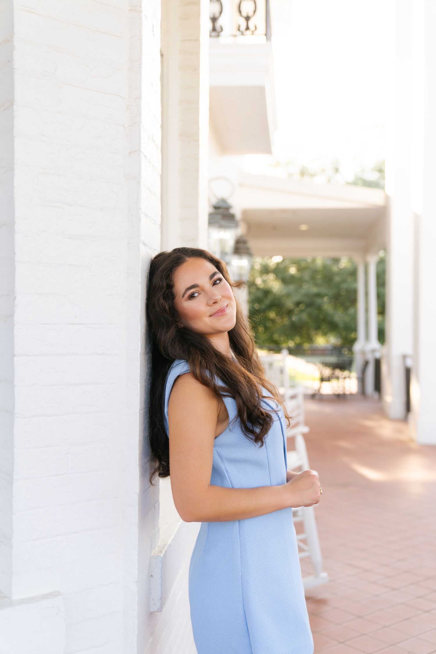 Someone in a light blue sleeveless dress poses against a white column on a brick porch in warm sunlight.