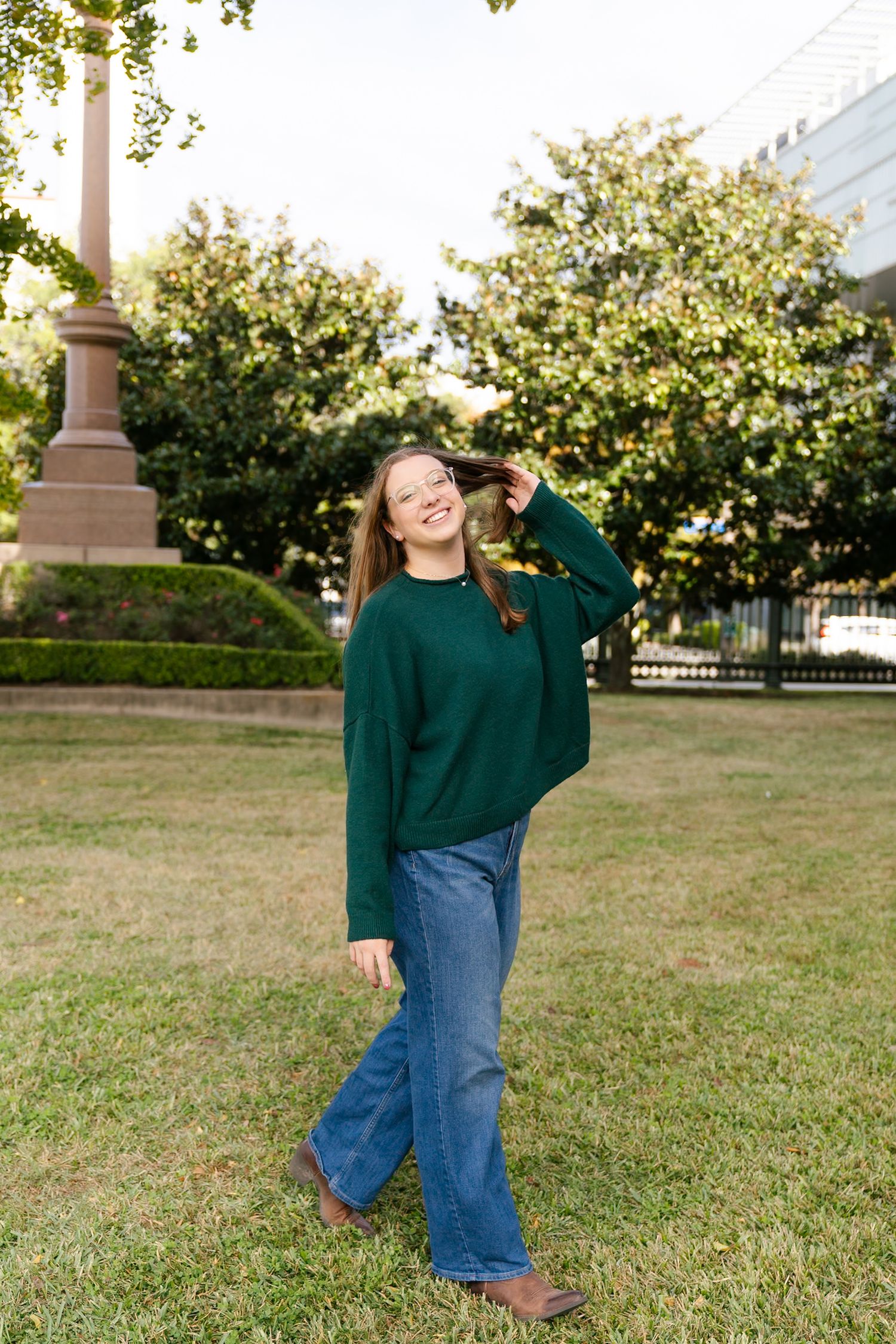 A person wearing a green sweater and blue jeans poses casually on grass near a lamppost in a park setting.