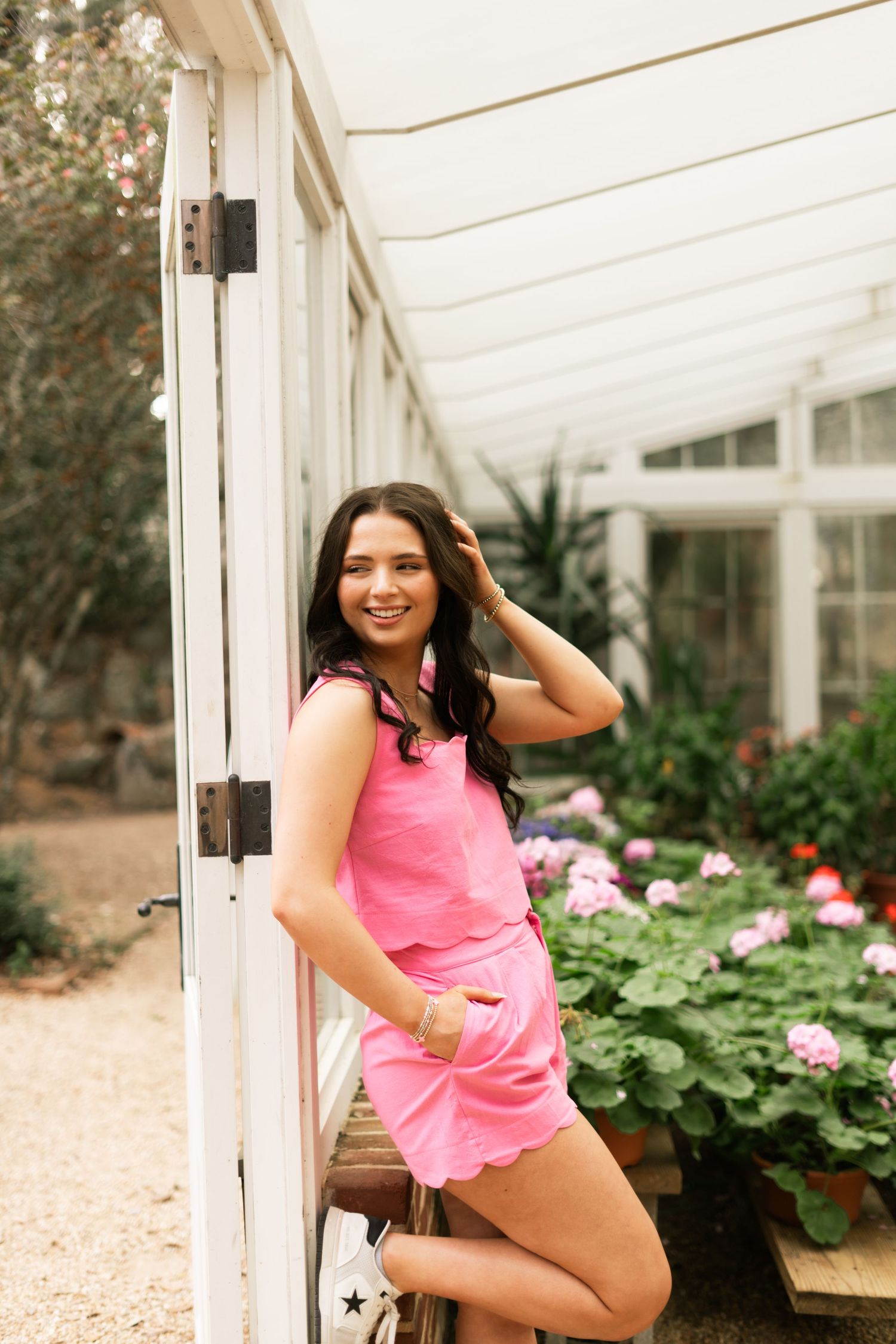 A person in a pink tank top poses casually against a white greenhouse wall surrounded by flowering plants.