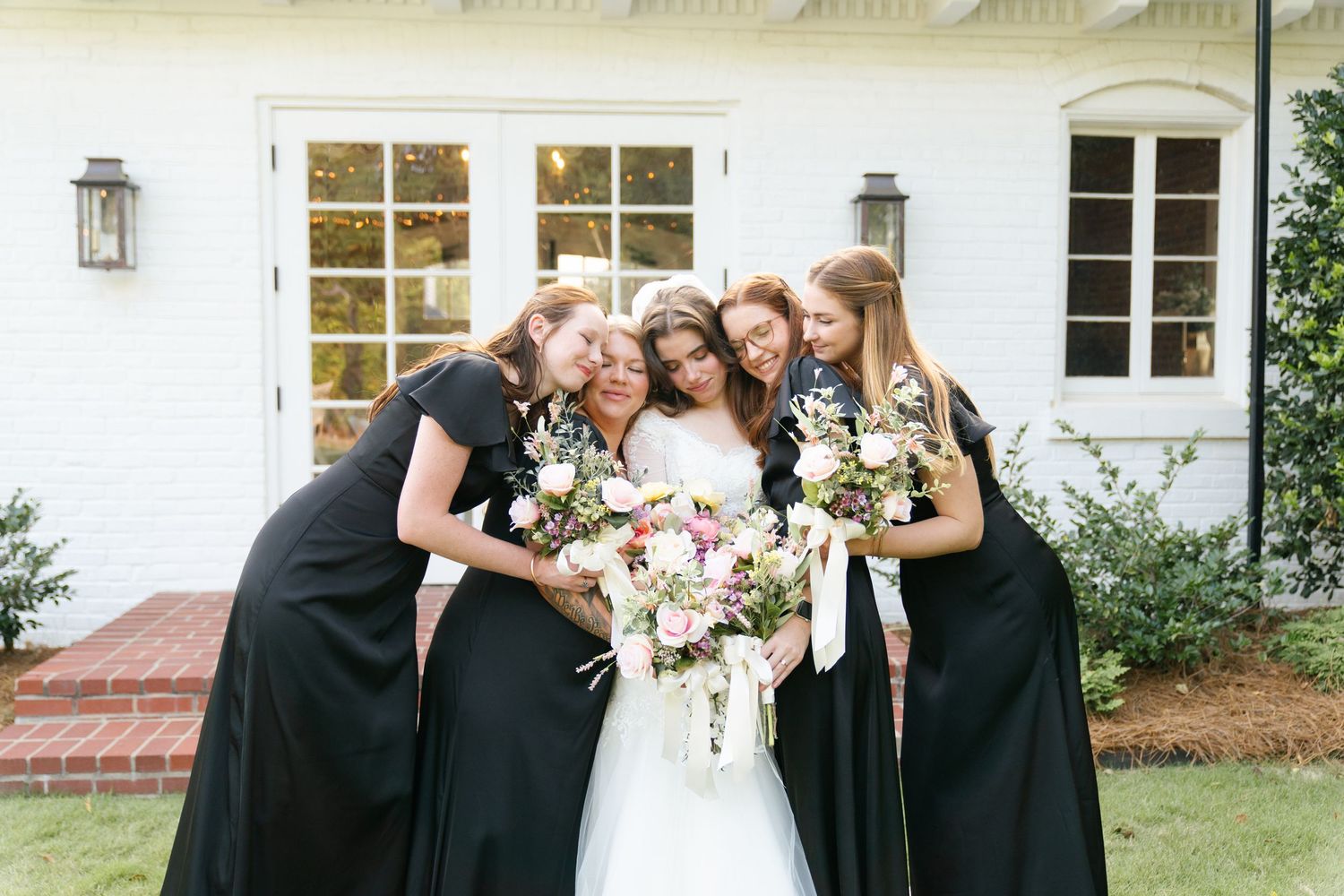 Group gathered outside white building dressed in formal attire holding bouquets.