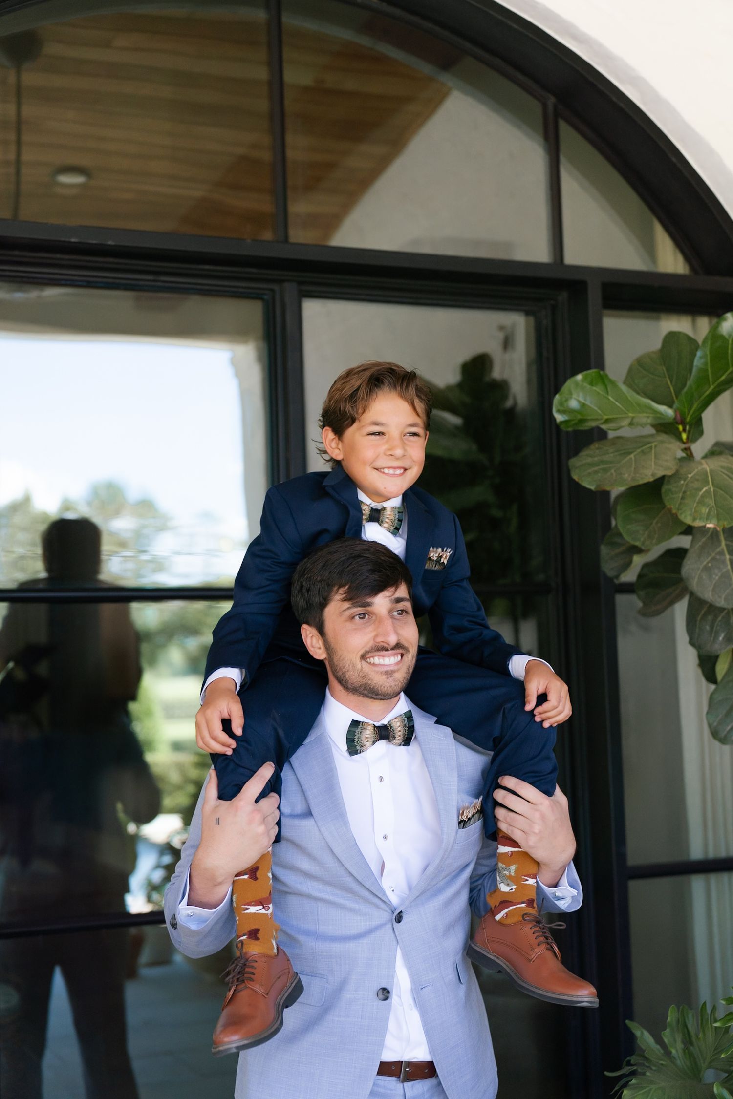 Groomsman in light gray suit playfully carries young ring bearer in navy suit during wedding photos.