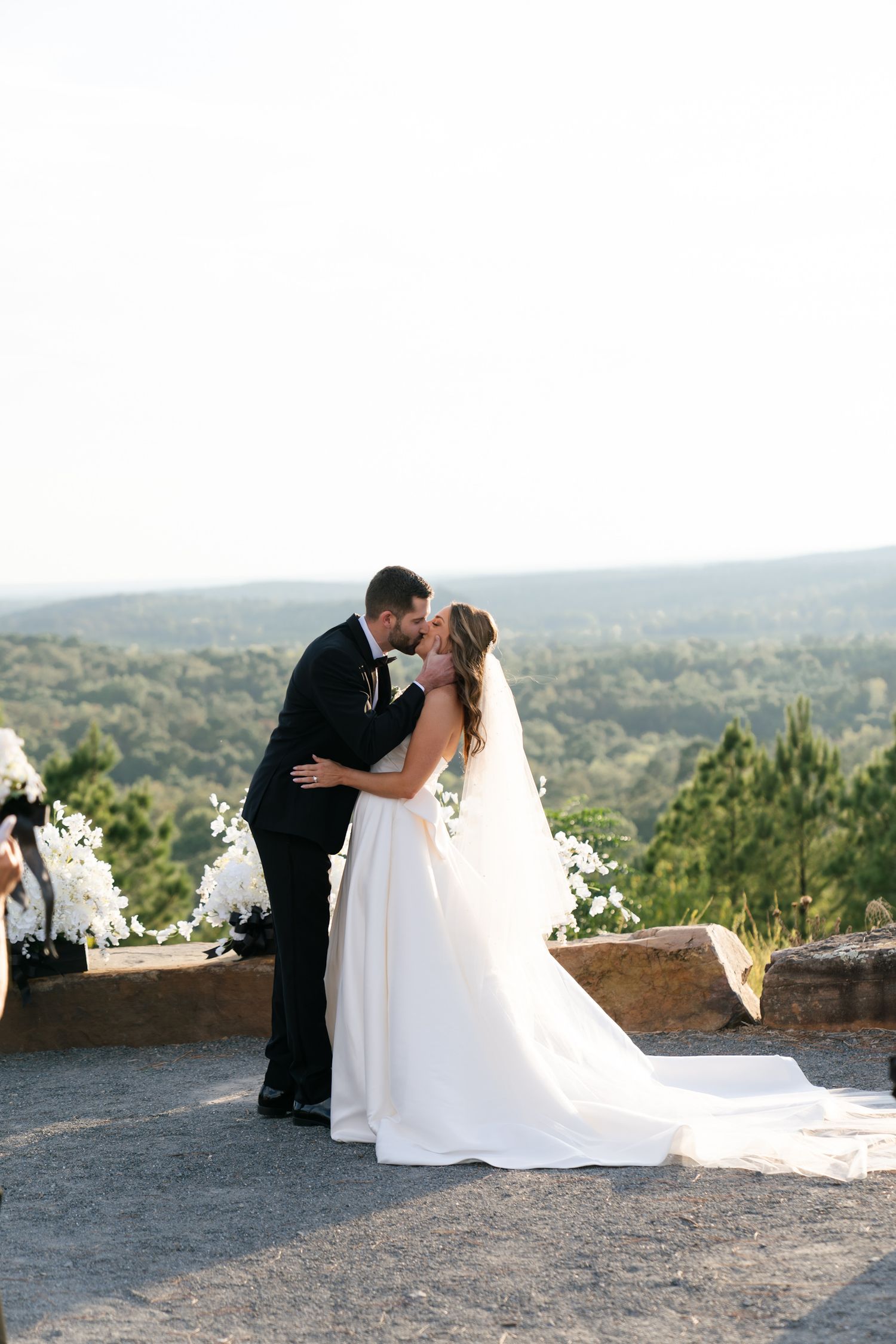 A bride and groom share a romantic kiss at a scenic mountain overlook during their outdoor wedding ceremony.