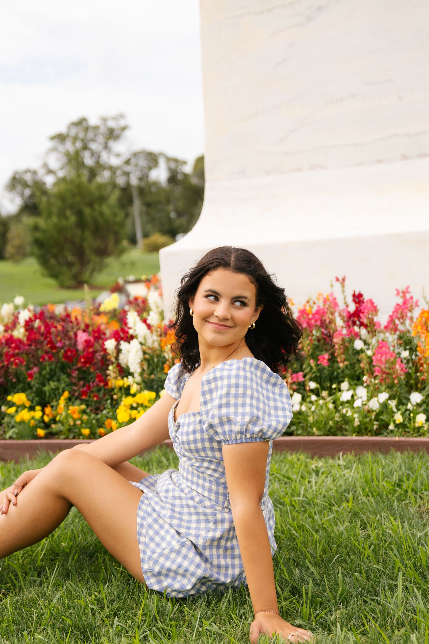 A person in a gingham dress poses in a garden with vibrant red and yellow flowers blooming in the background.