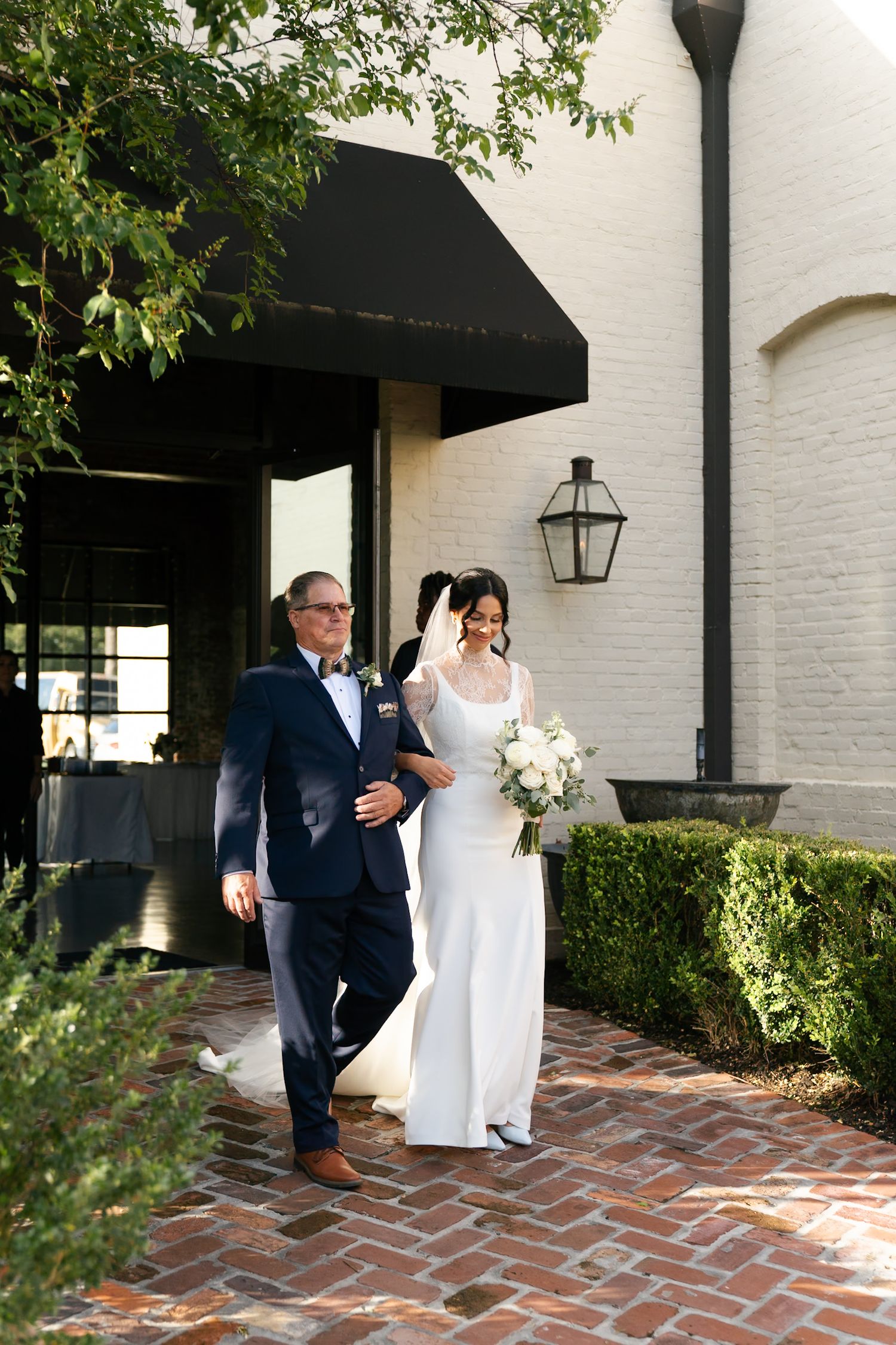 Wedding couple walks along brick pathway outside white building with black accents.