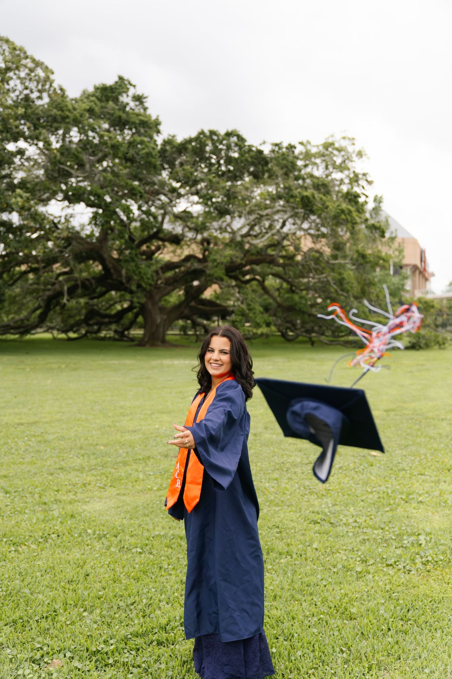 Graduate in blue cap and gown celebrates on green lawn with orange stole.