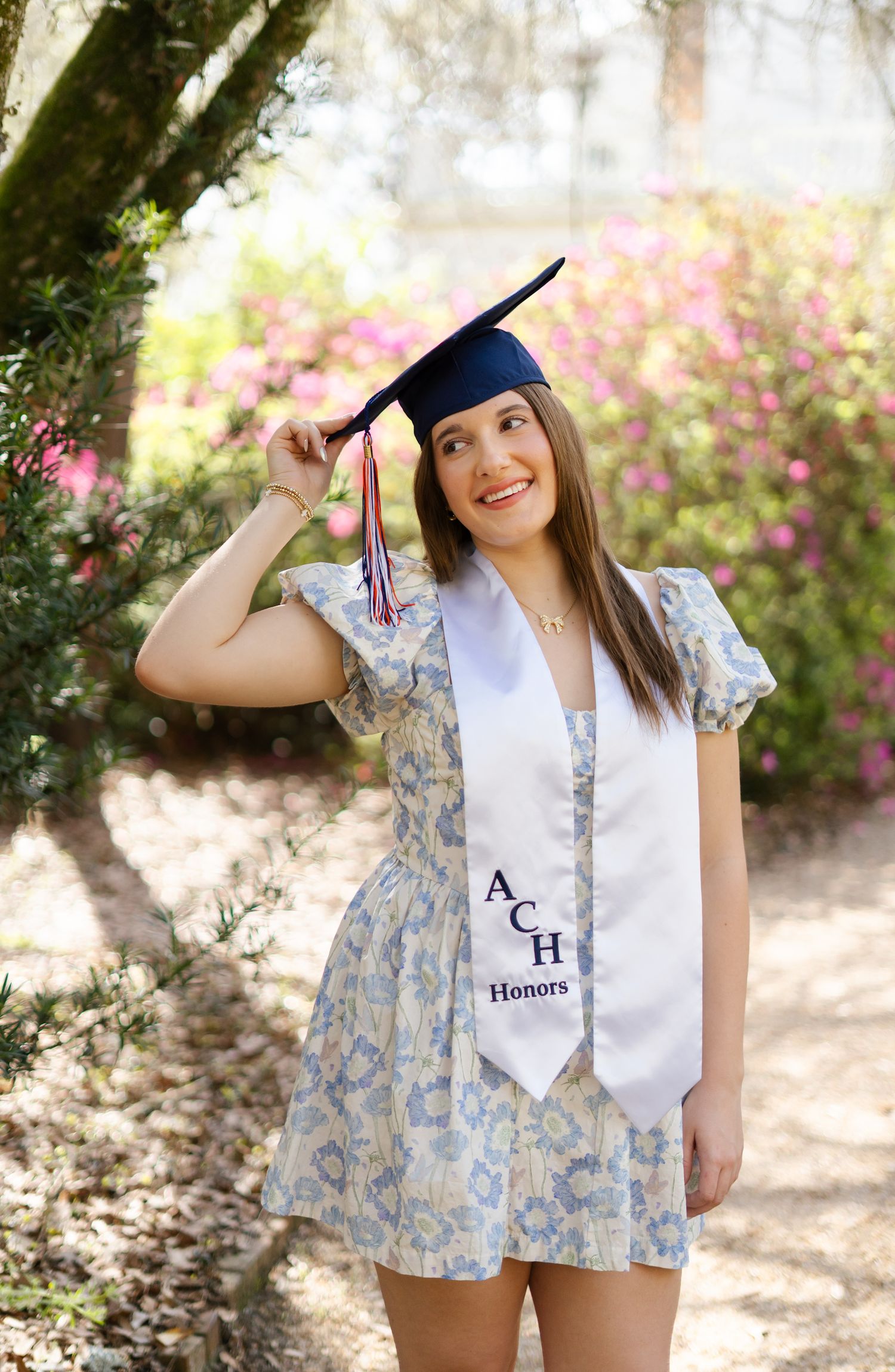 Graduate wearing white stole and graduation cap poses outdoors with pink flowers in the background.