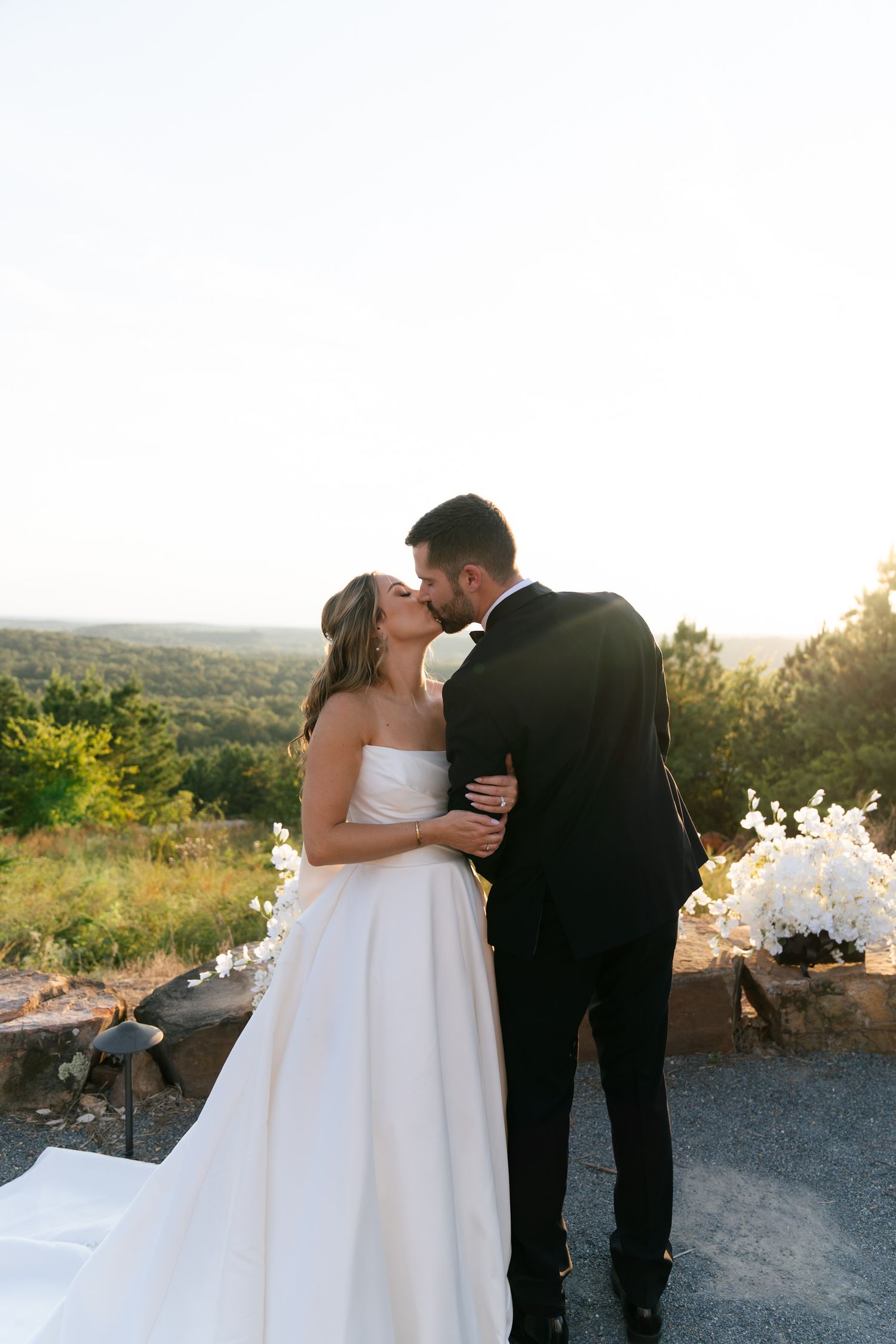 A romantic couple in wedding attire share intimate moments at sunset on a hilltop overlooking a scenic landscape.