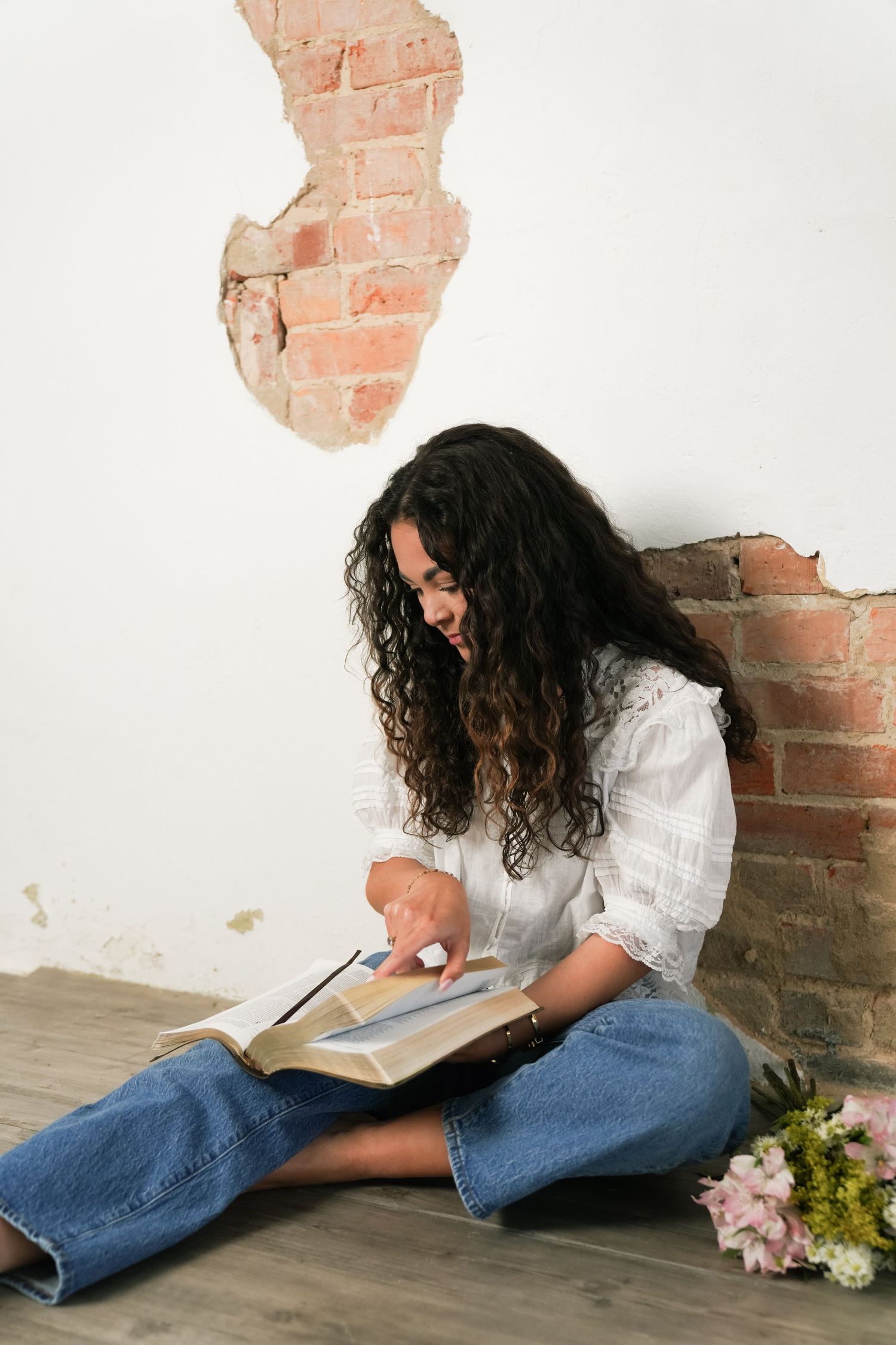 A person in jeans and white shirt lies on floor reading a book beside exposed brick wall and flowers.
