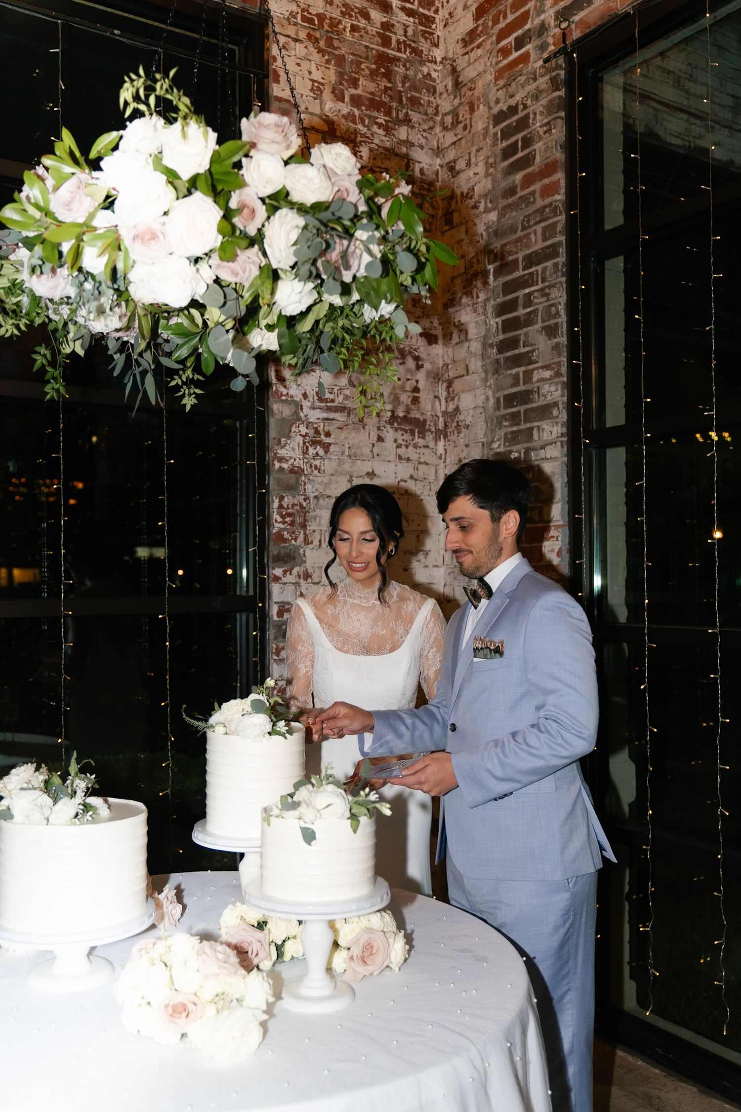 Couple cuts their white wedding cake together against exposed brick wall with hanging lights.