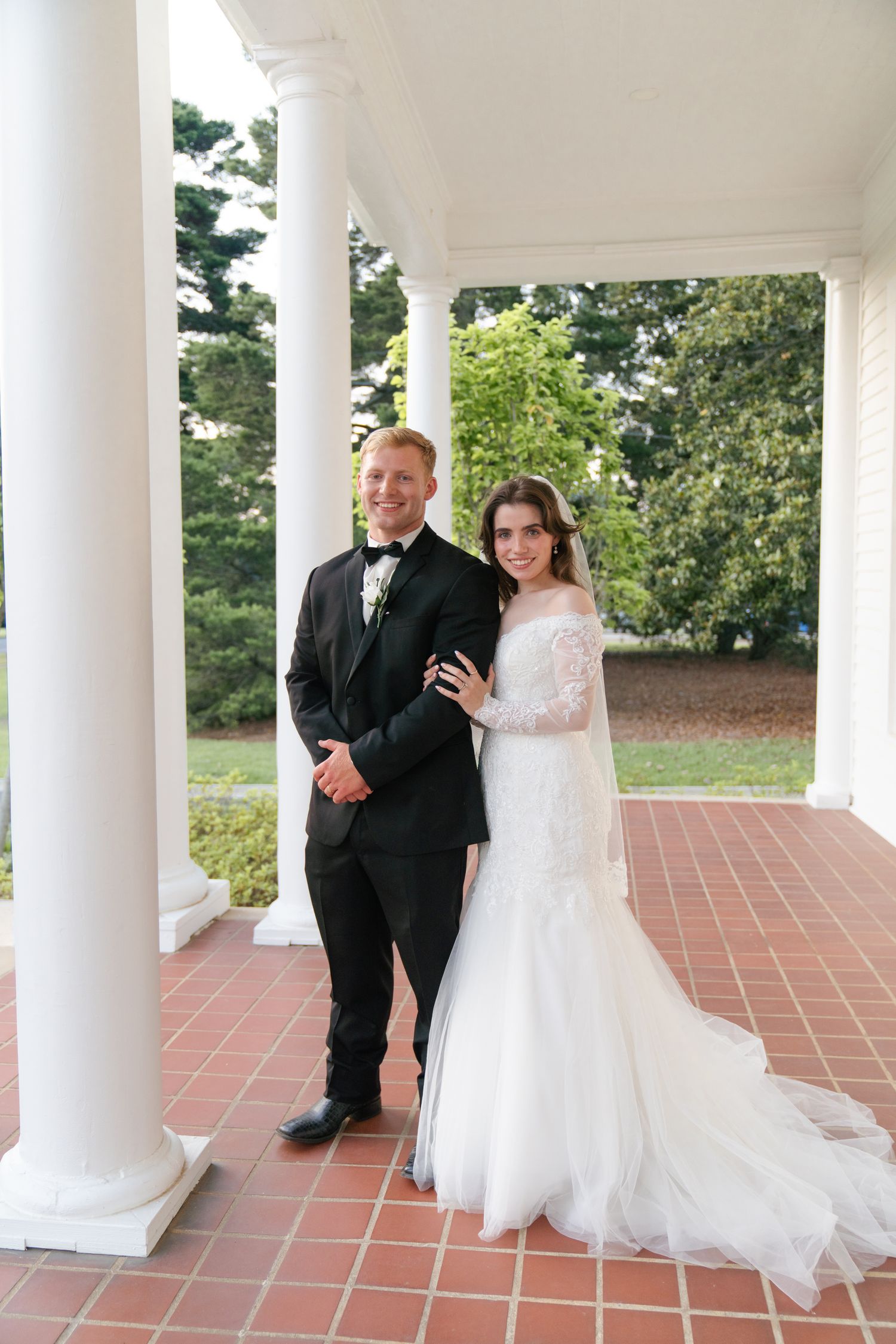 Wedding party gathered under white columned porch with red brick flooring.