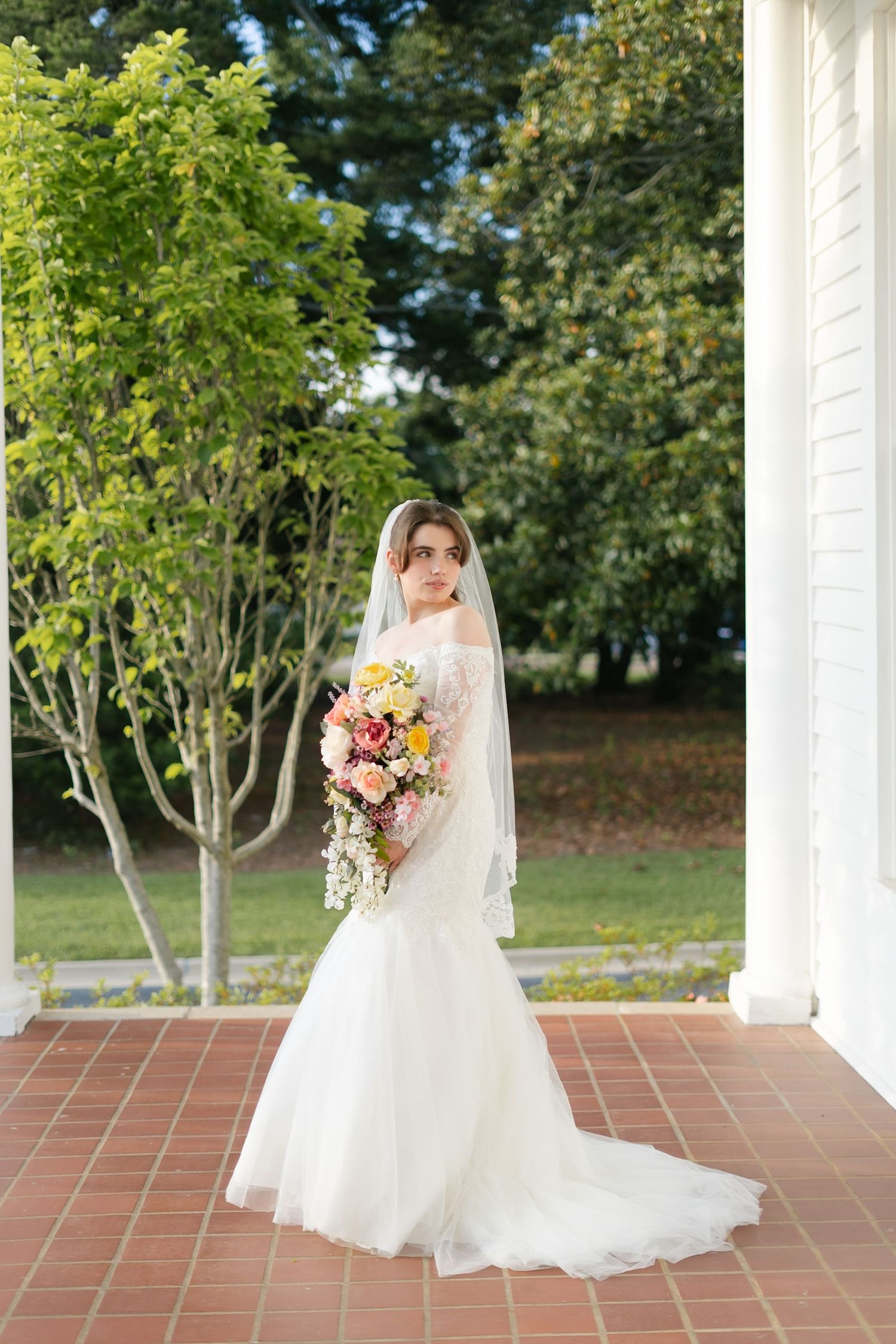 Person in wedding dress holding bouquet under classic white colonnade.